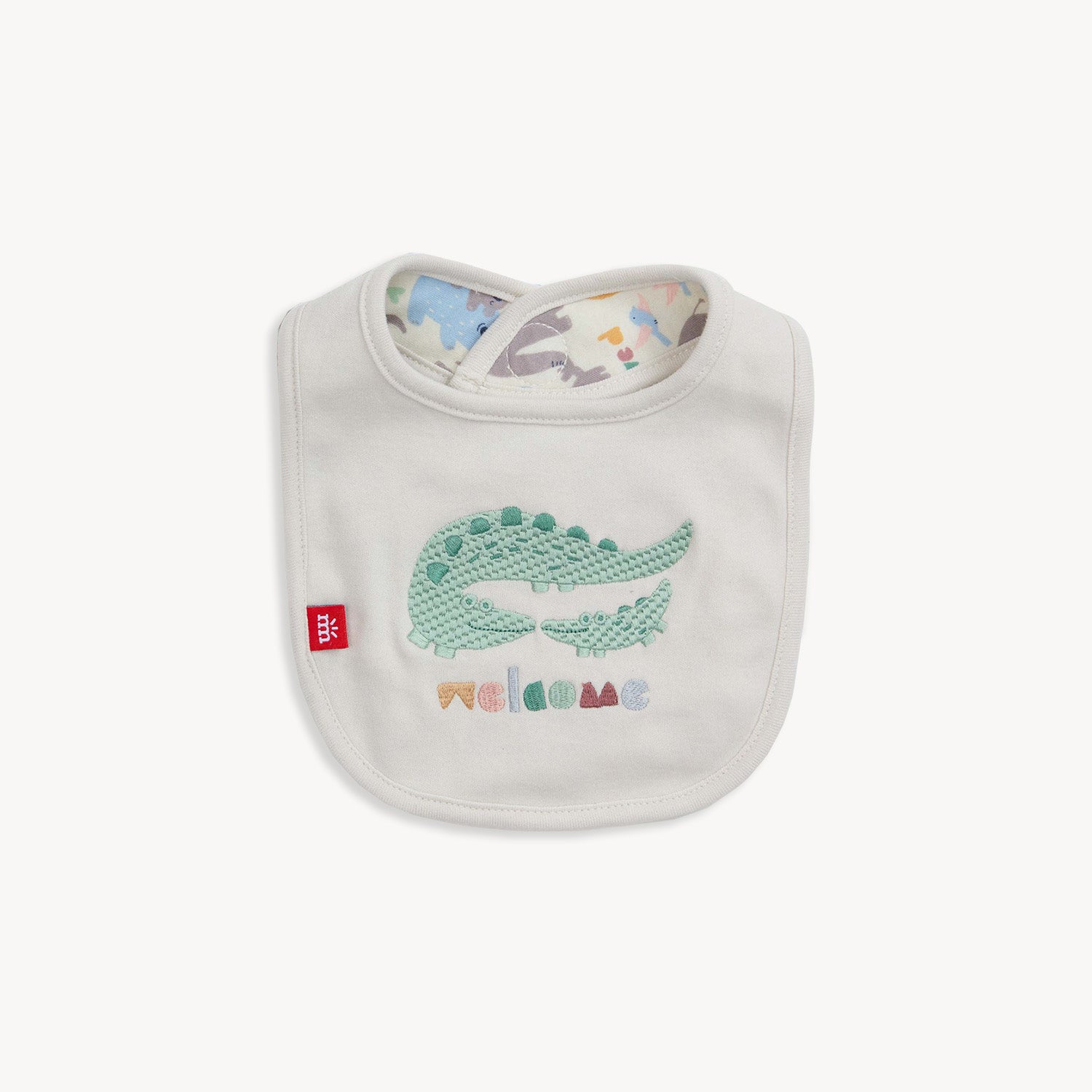 A cream-colored reversible infant bib with a green crocodile and "welcome" embroidery hanging from a clothesline  by Magnetic Me.