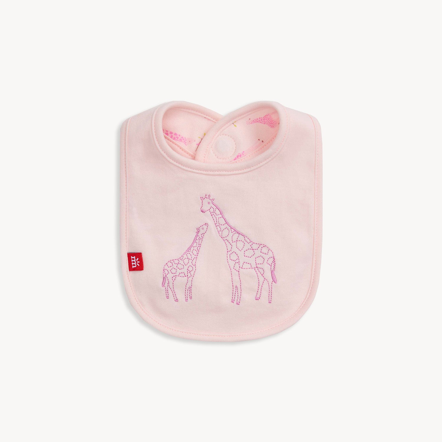 A light pink reversible bib featuring a large embroidered giraffe and baby giraffe on a solid background, with a repeat Jolie Giraffe print on the reverse side, by Magnetic Me.
