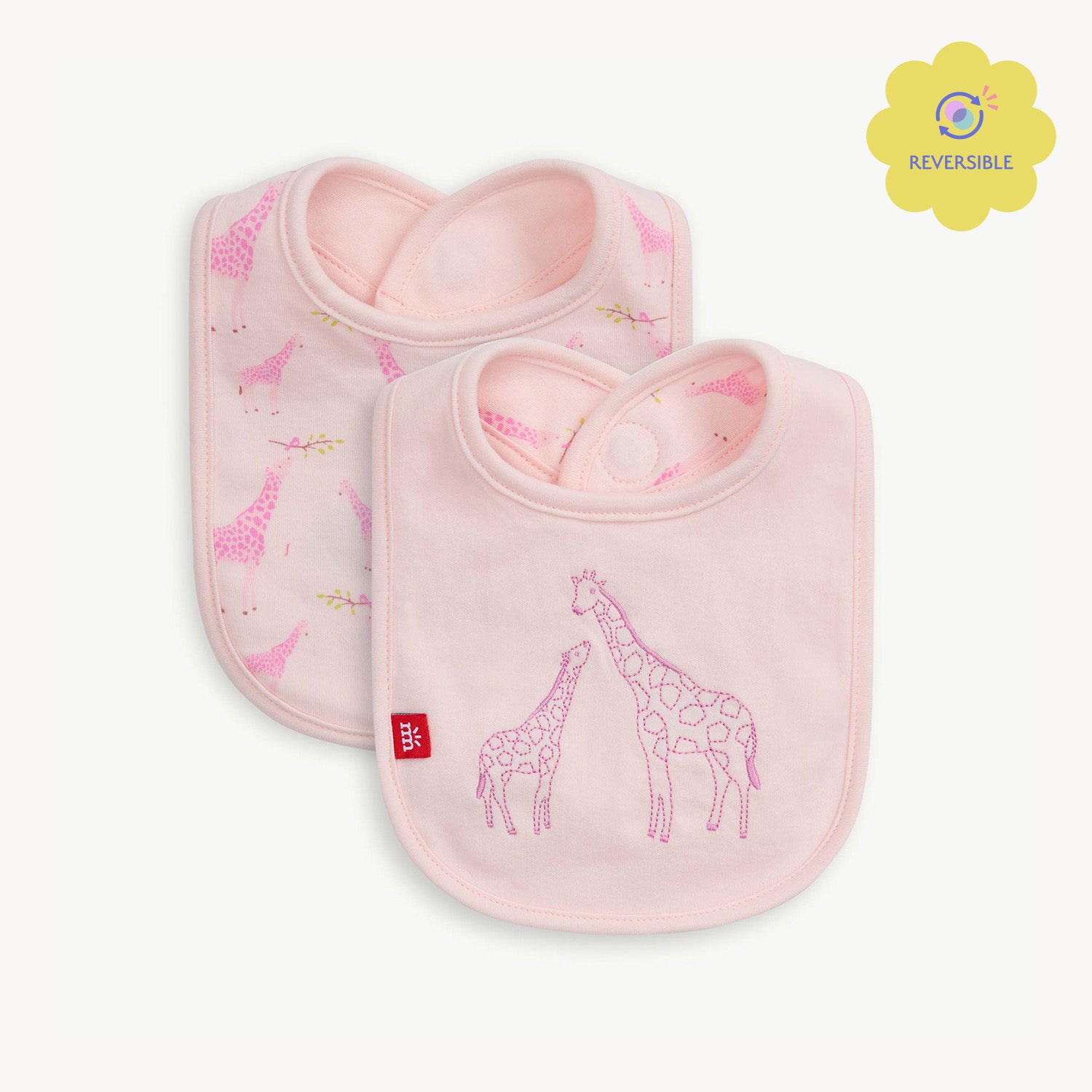 A light pink reversible bib featuring a large embroidered giraffe and baby giraffe on a solid background, with a repeat Jolie Giraffe print on the reverse side, by Magnetic Me.