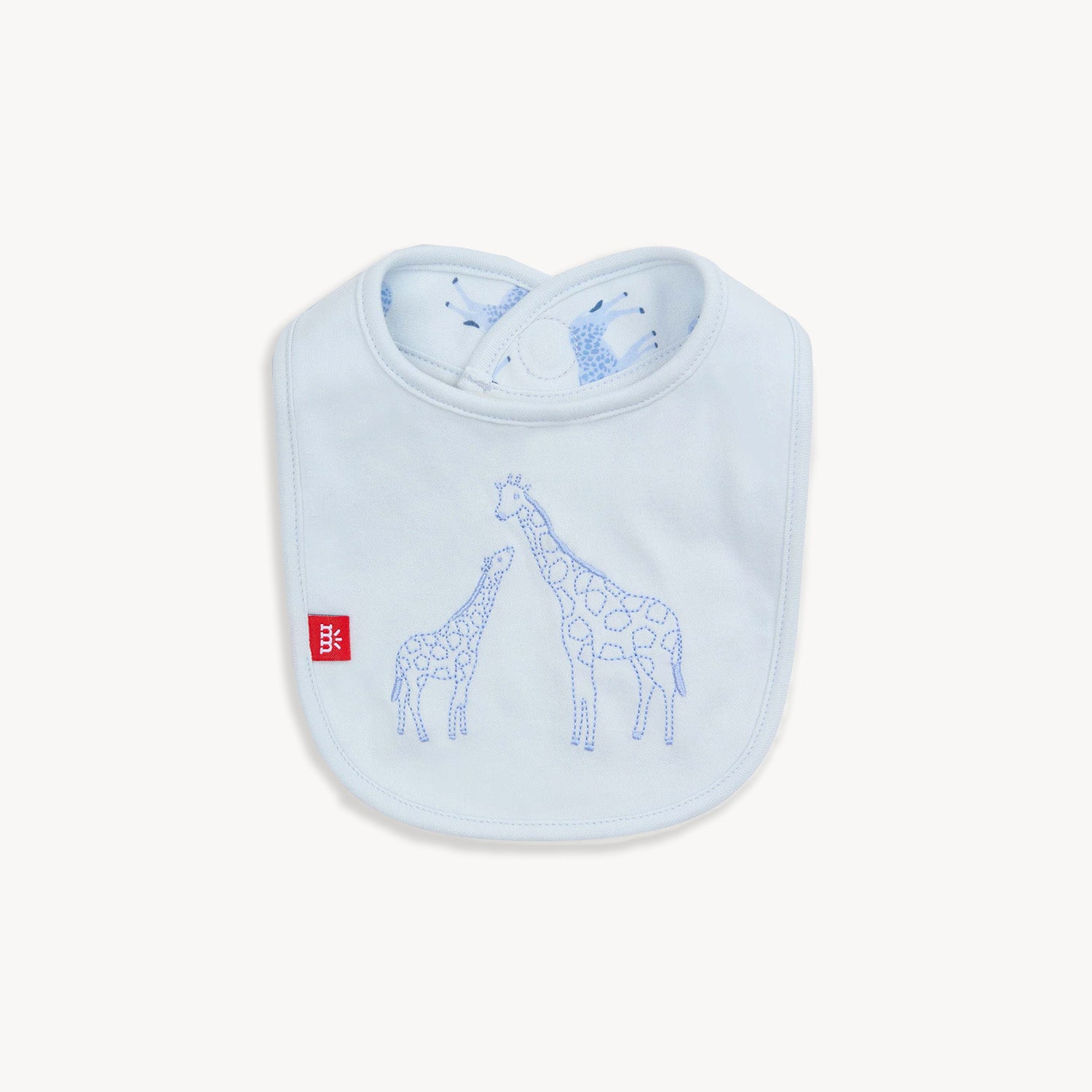 A pale blue reversible bib featuring a large embroidered giraffe and baby giraffe on a solid background, with a repeat Jolie Giraffe print on the reverse side, by Magnetic Me.