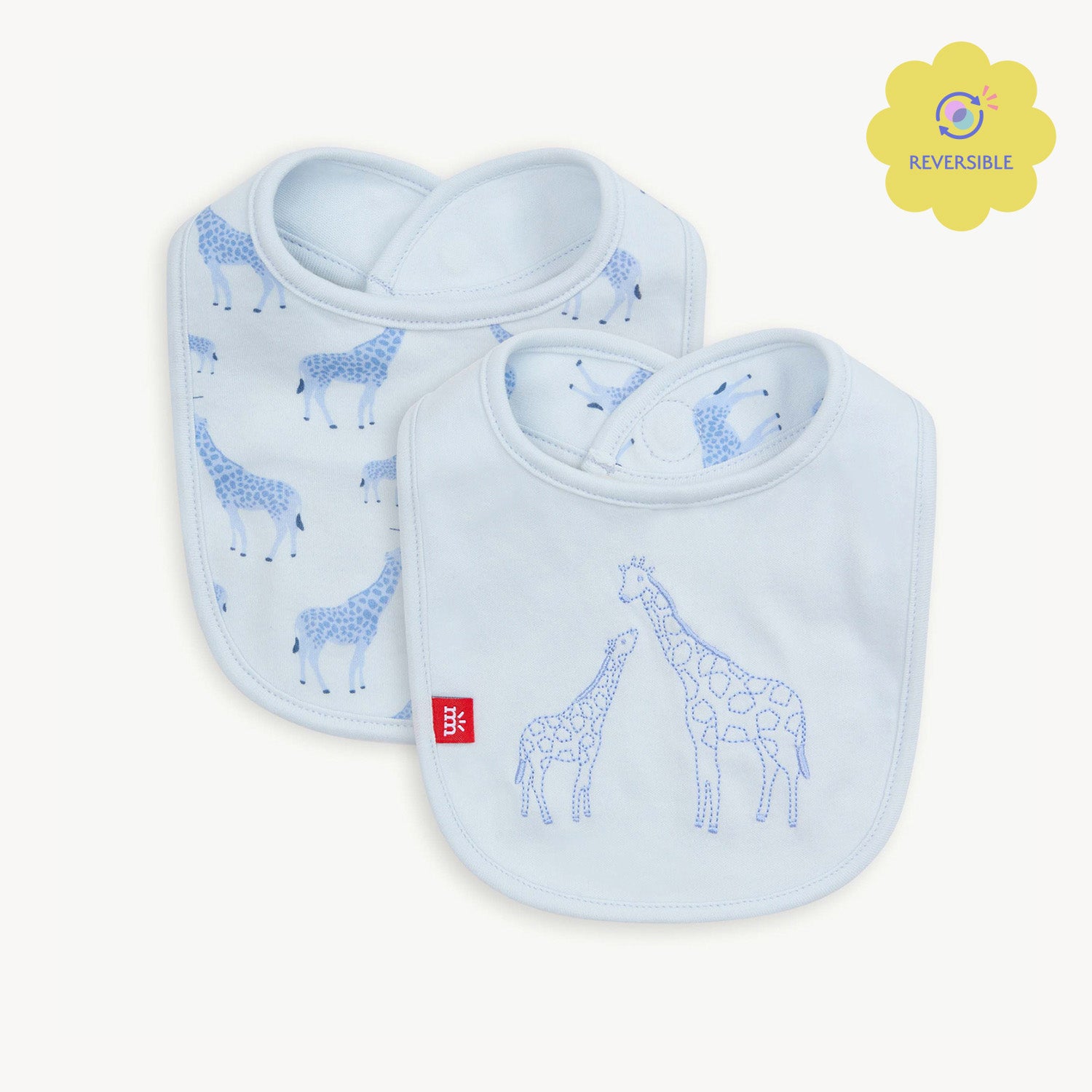 A pale blue reversible bib featuring a large embroidered giraffe and baby giraffe on a solid background, with a repeat Jolie Giraffe print on the reverse side, by Magnetic Me.