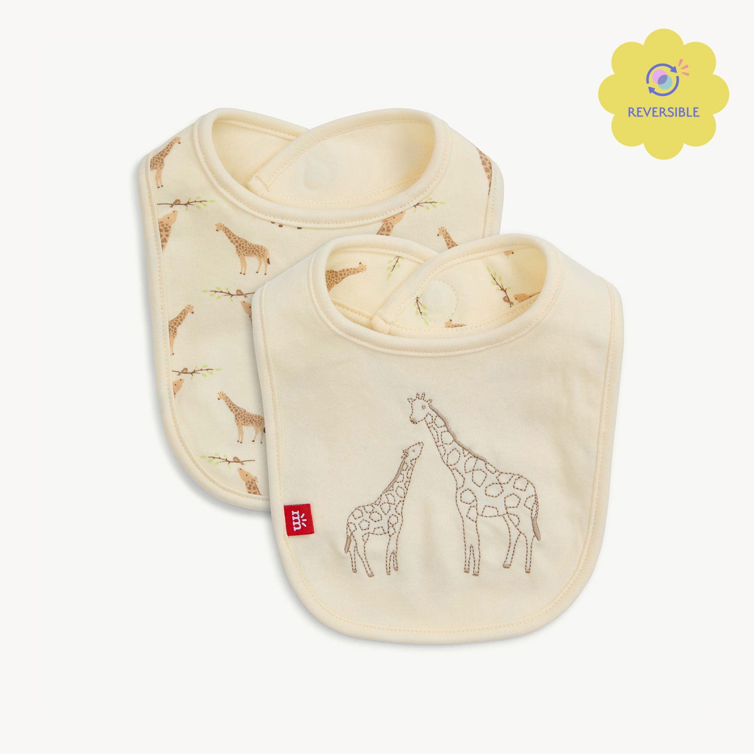 A cream-colored reversible bib featuring a large embroidered giraffe and baby giraffe on a solid background, with a repeat Jolie Giraffe print on the reverse side, by Magnetic Me.