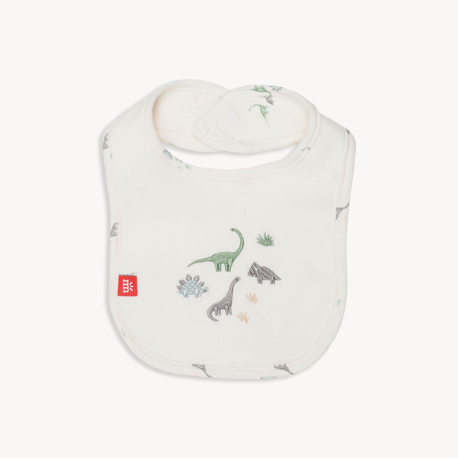 reversible cream-colored bib featuring a watercolor dinosaur print on one side and a larger embroidery of three different dinosaurs on the other, by Magnetic Me.