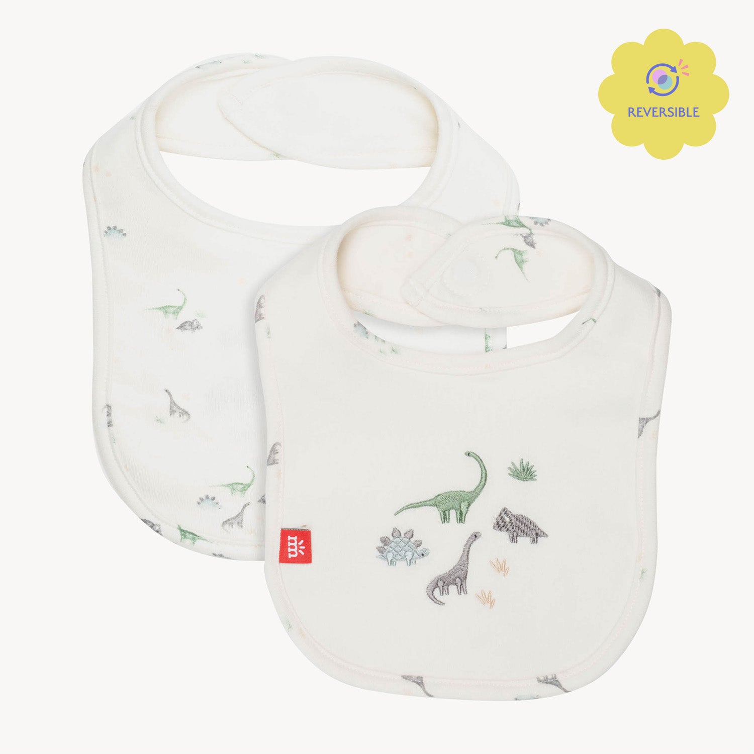 reversible cream-colored bib featuring a watercolor dinosaur print on one side and a larger embroidery of three different dinosaurs on the other, by Magnetic Me.