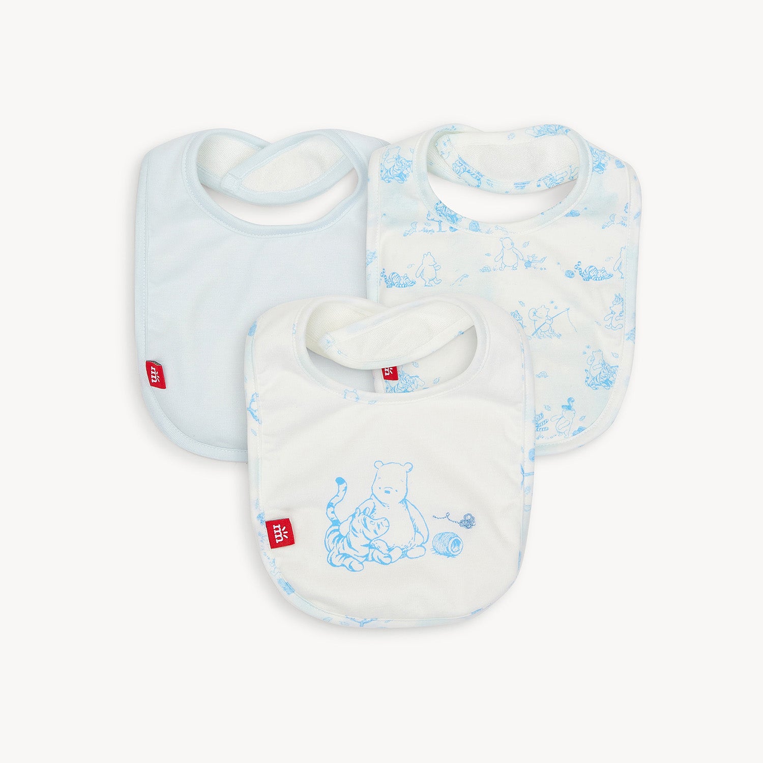 Set of three infant bibs in light blue modal fabric featuring a Disney Winnie the Pooh "In The Woods" blue sketch print, including one solid light blue bib, one with an all-over character sketch print, and one with a central illustration of Winnie the Pooh and Tigger with a ball of yarn, by Magnetic Me.
