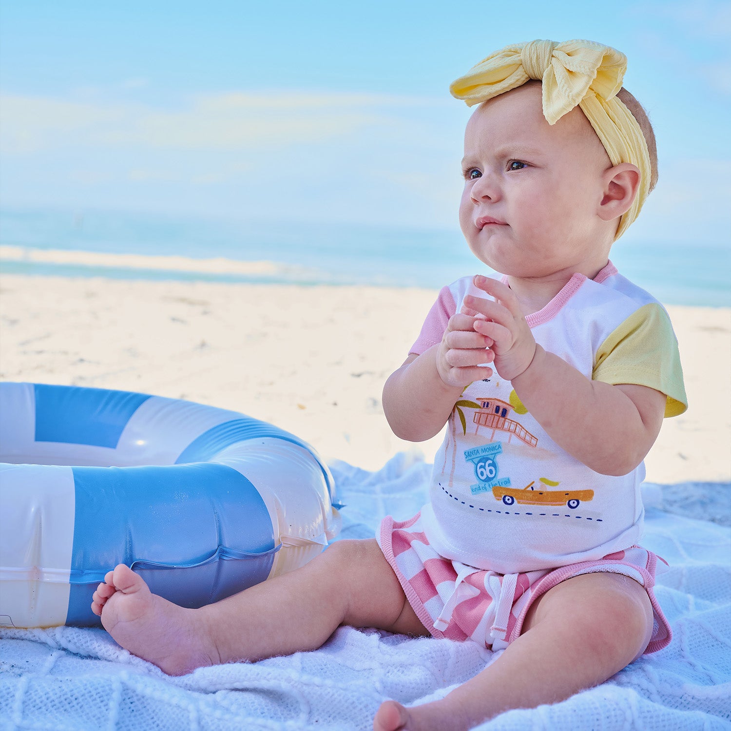 Baby sitting on a towel at the beach with a life buoy in the background wearing A retro-inspired outfit features a white short-sleeve tee with one pink and one yellow sleeve, a Santa Monica beach scene graphic, and coordinating pink-and-white vertical striped shorts by Magnetic Me.