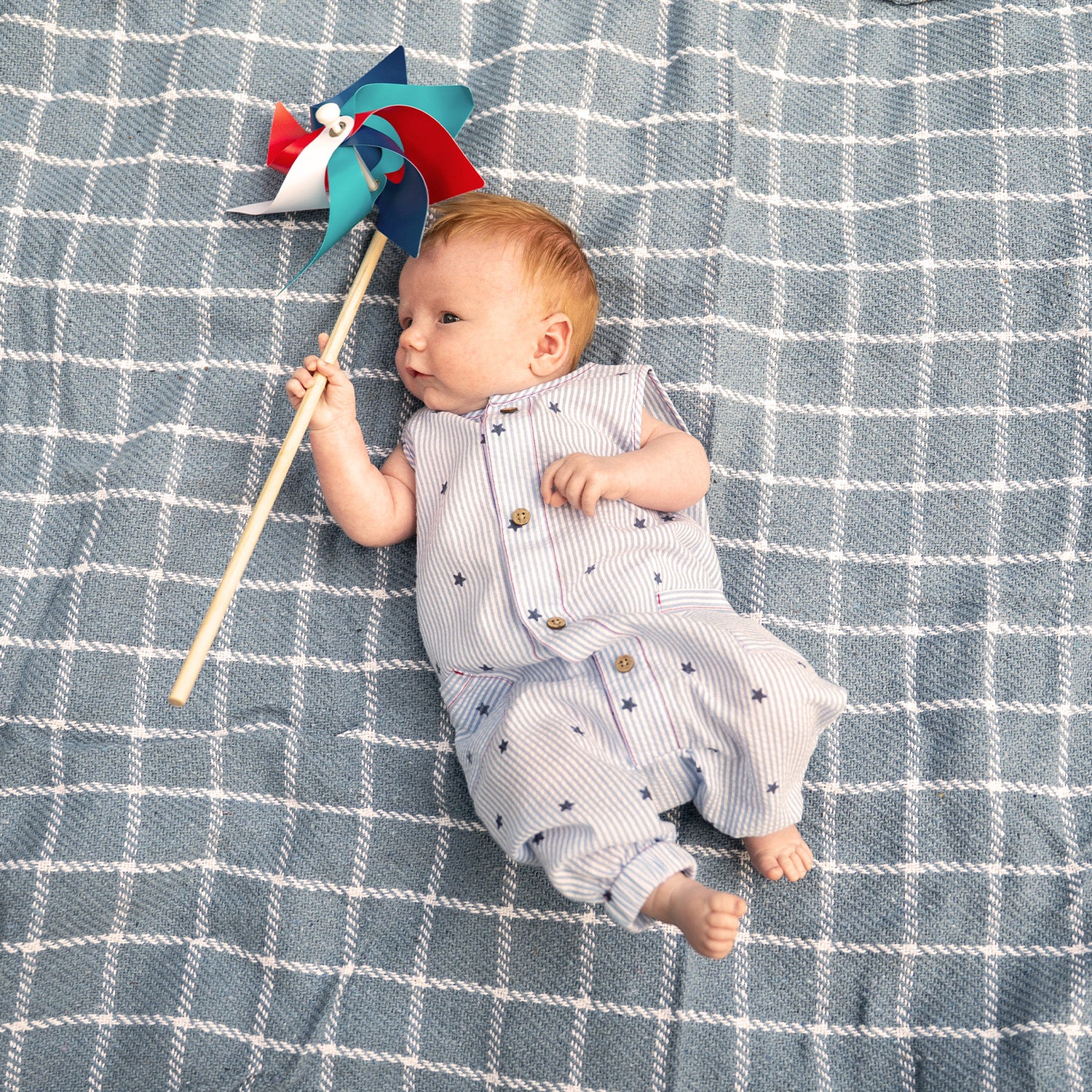 Baby lying on a checkered blanket holding a colorful pinwheel wearing A light blue and white striped seersucker sleeveless romper featuring navy star print, wood-look buttons, and front pockets, by Magnetic Me.