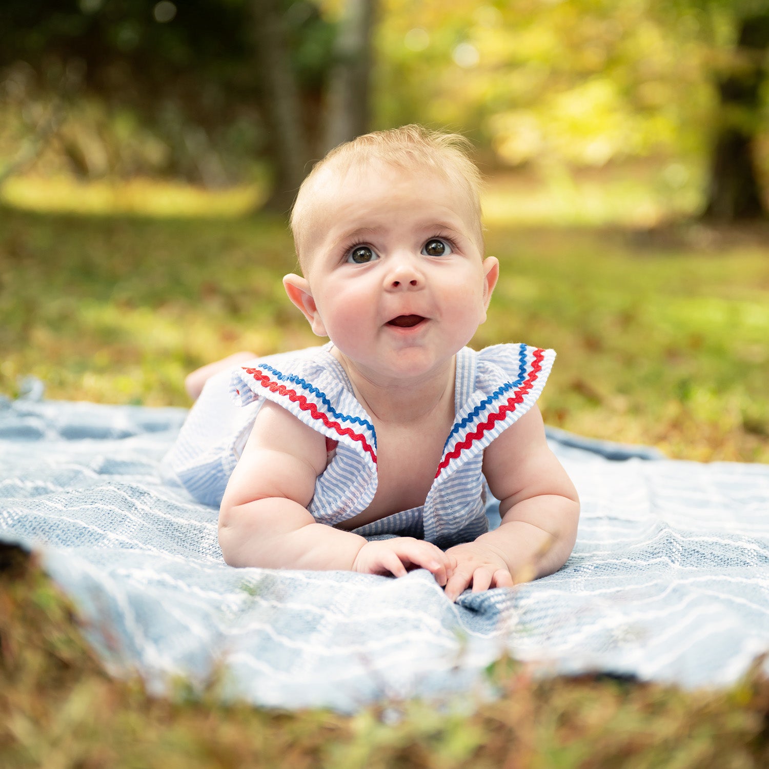 Baby lying on a blanket in a park with a blurred natural background wearing A light blue striped seersucker bubble romper featuring ruffled shoulder straps accented with red and blue rickrack trim, by Magnetic Me.
