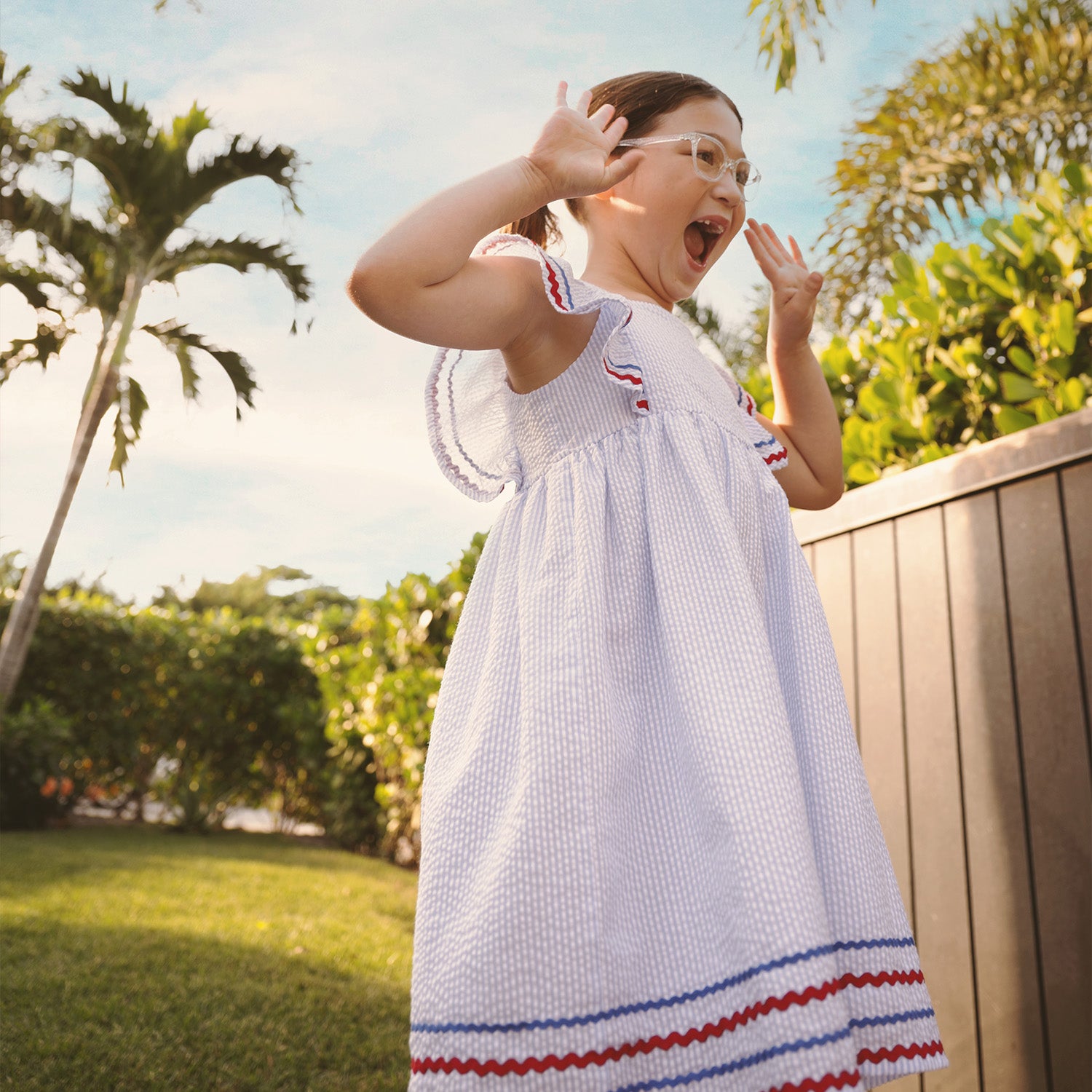 Child standing outdoors with trees and a wooden fence in the background wearing A light blue striped seersucker sleeveless dress featuring ruffled straps and a tiered hem detailed with red and blue rickrack trim, by Magnetic Me.