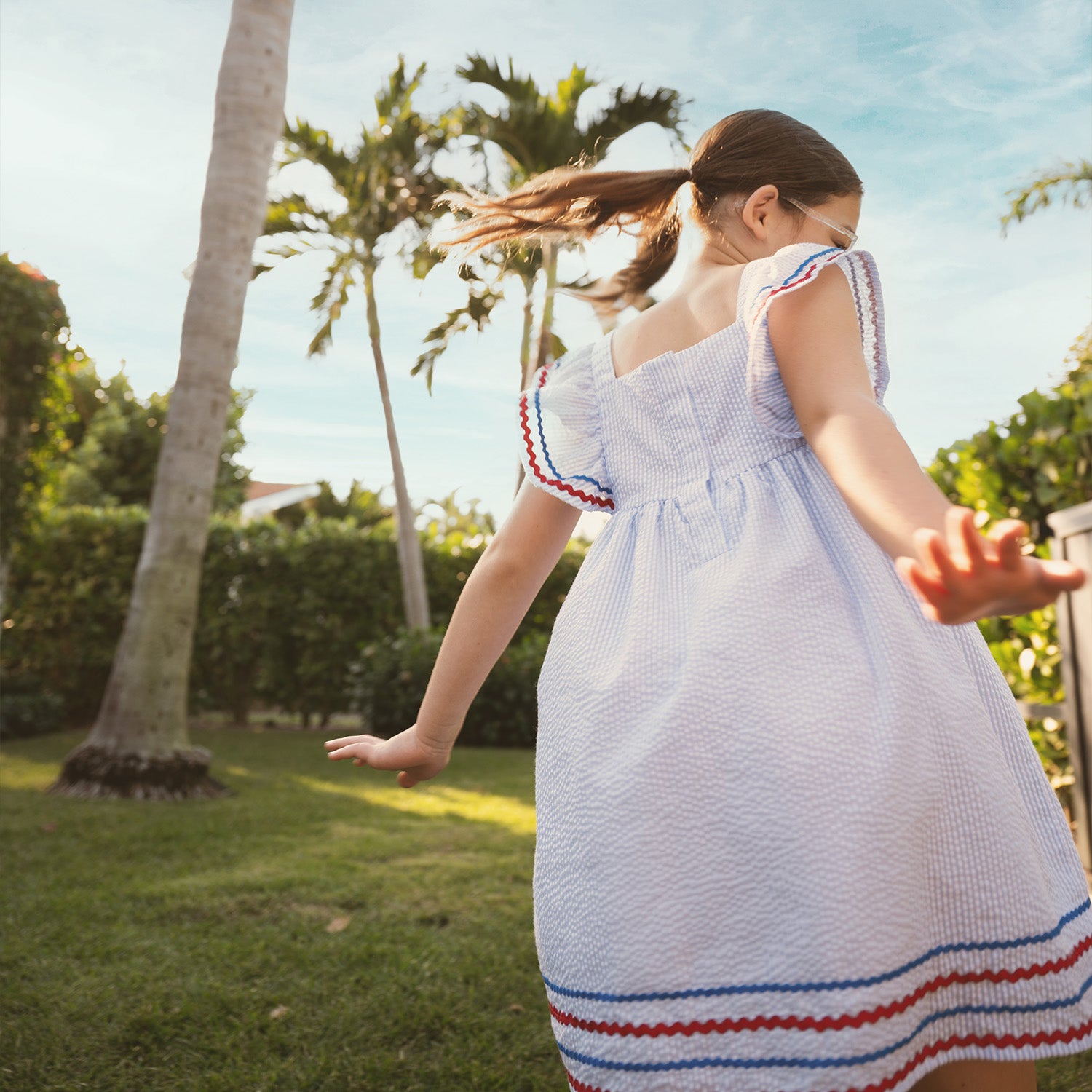 A young girl wearing A light blue striped seersucker sleeveless dress featuring ruffled straps and a tiered hem detailed with red and blue rickrack trim, by Magnetic Me.