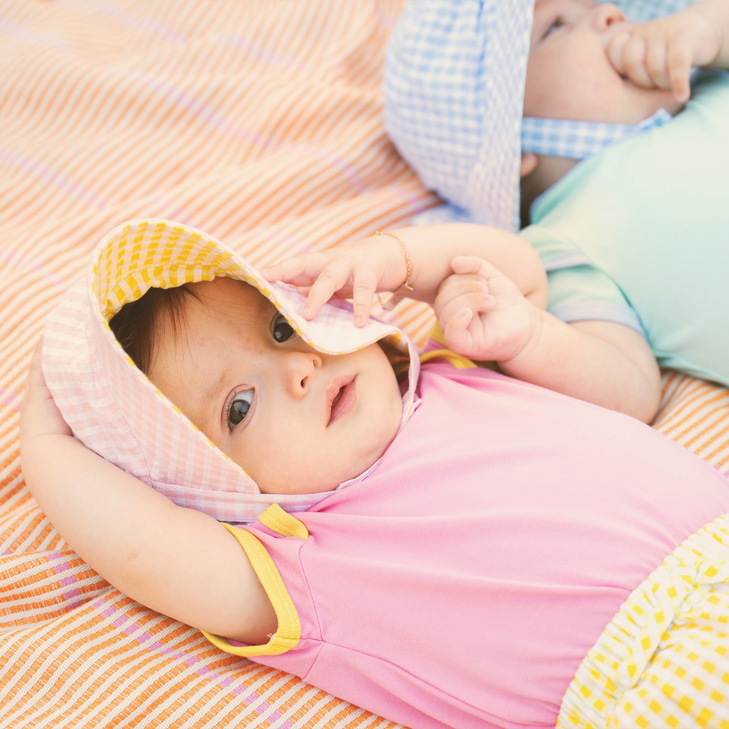 Two babies lying on a colorful blanket, one wearing a pink outfit with a yellow hat and the other in a blue outfit by Magnetic Me.