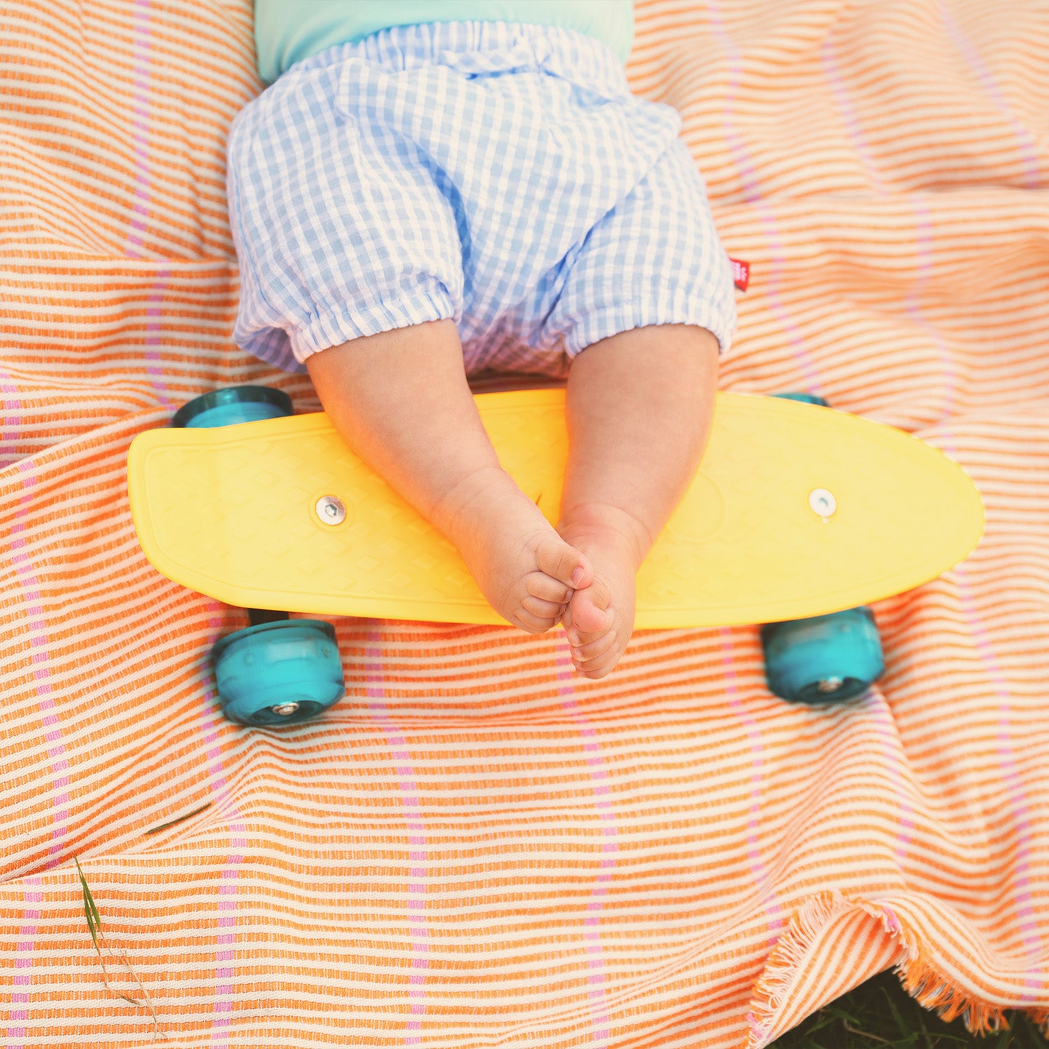 Child's legs on a yellow skateboard with blue wheels on an orange striped blanket wearing A two-piece set featuring a light green short-sleeve bodysuit with blue trim and matching blue gingham bloomers with a functional back pocket, by Magnetic Me.