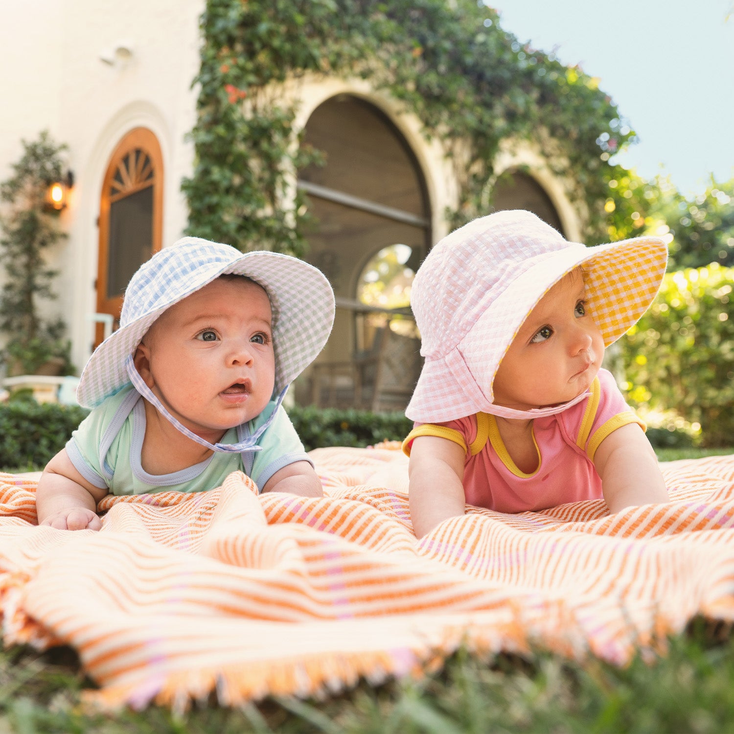 Two babies wearing bucket hats sitting on a striped blanket outdoors by Magnet Me.