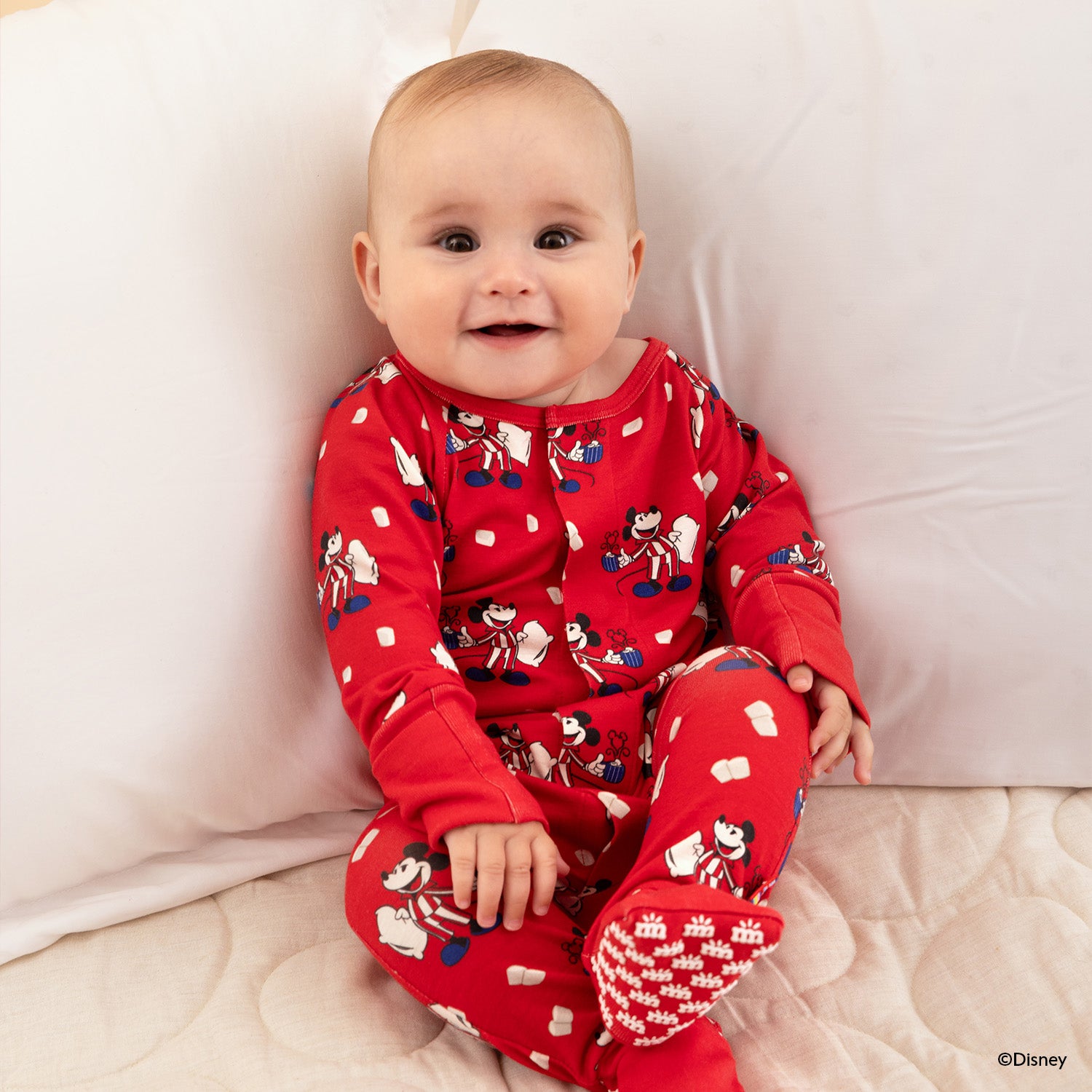 Baby sitting and wearing A vibrant red convertible coverall featuring a Disney-themed print of Mickey Mouse in striped pajamas holding a mug and pillow, accented with floating marshmallow motifs and a concealed magnetic closure for quick dressing by Magnetic Me.