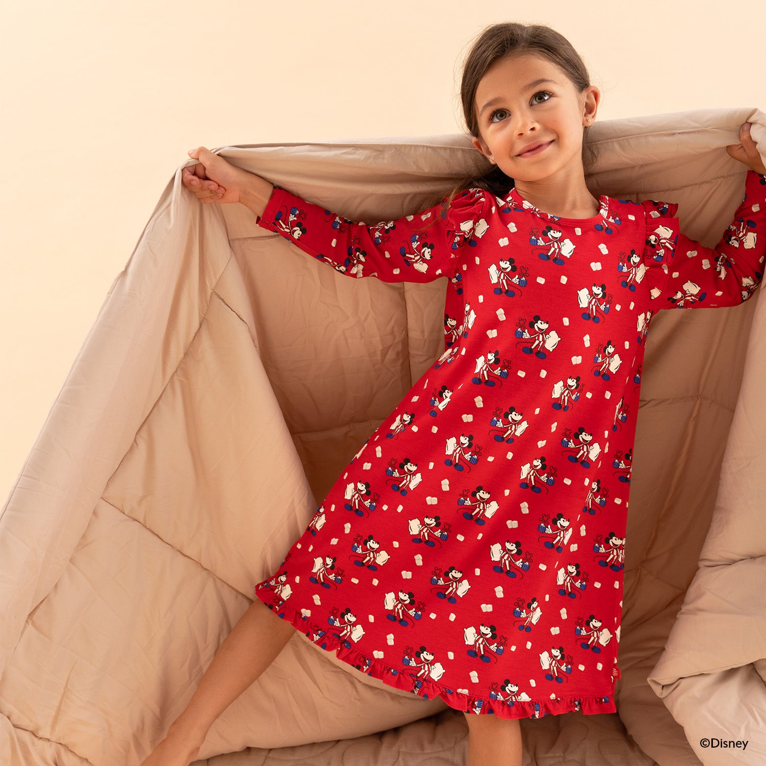 Young girl wearing A vibrant red long-sleeve nightgown featuring a Disney print of Mickey Mouse in striped pajamas holding a mug and pillow, detailed with ruffle trim at the shoulders and hem by Magnetic Me.
