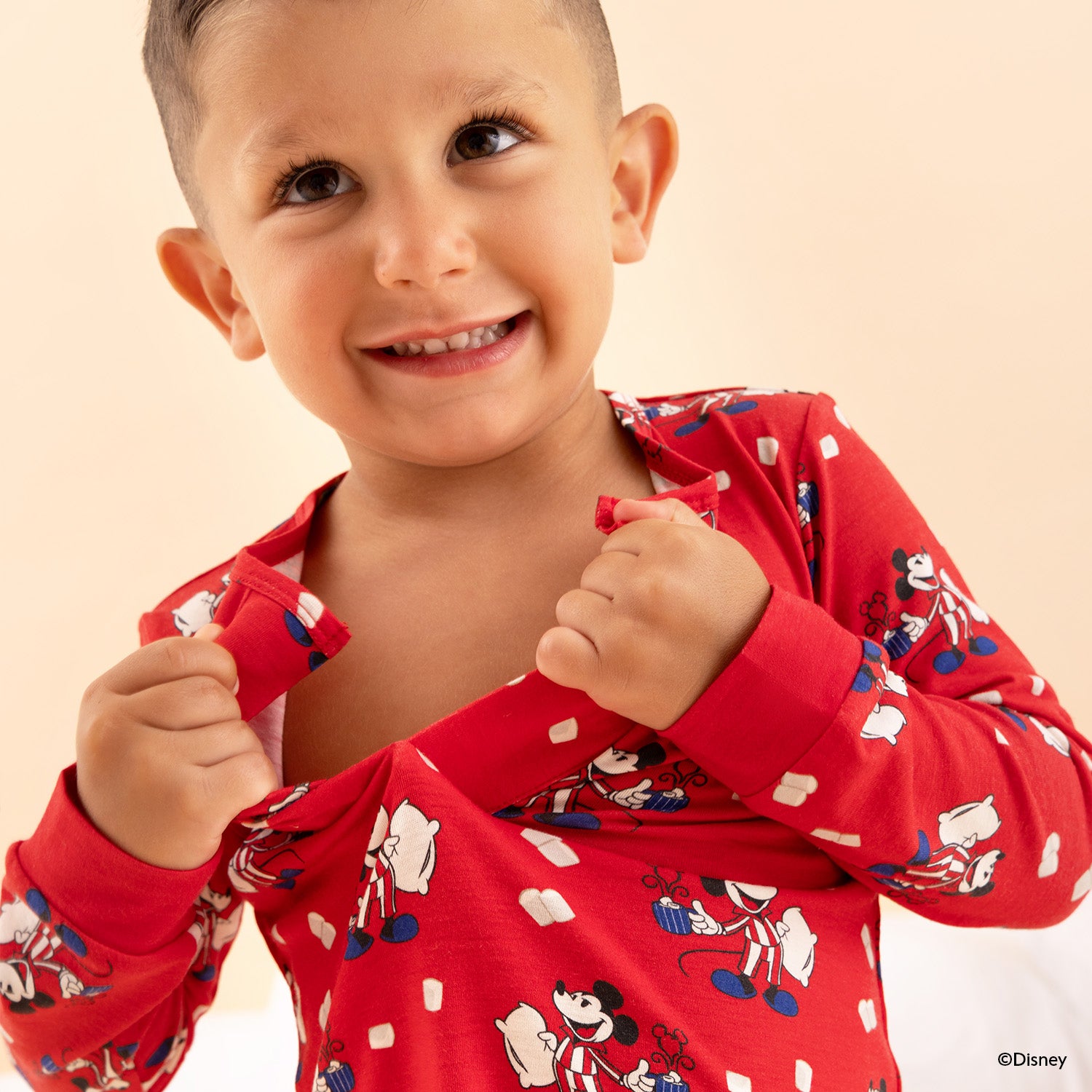 Boy opening the magnet in the front of A two-piece pajama set in red fabric featuring a long-sleeve top with hidden magnets and matching joggers, both detailed with an all-over print of Mickey Mouse holding pillows, by Magnetic Me.