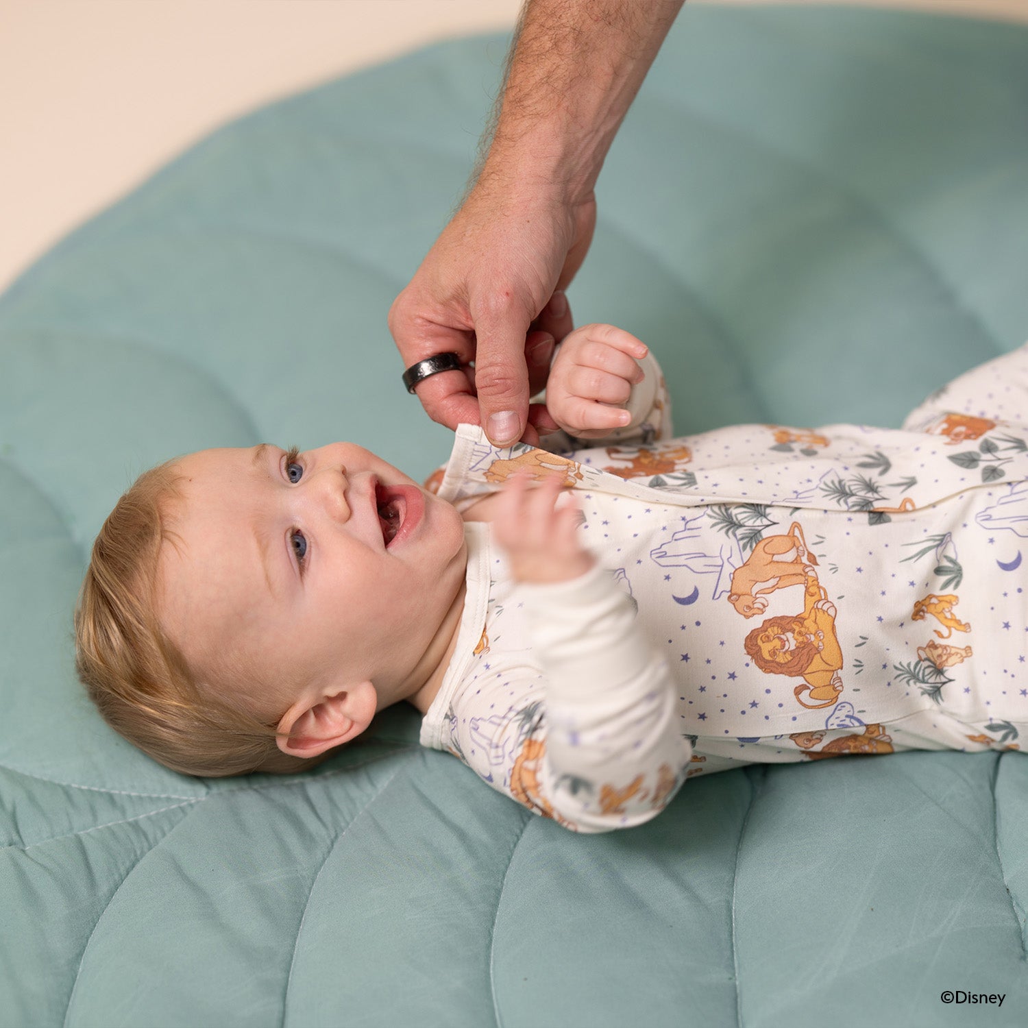 Baby lying on a green cushion with a person helping to adjust the magnet of A cream-colored long-sleeve coverall featuring a repeating pattern of Disney Pumbaa, Simba, and Nala among savanna trees and blue stars by Magnetic Me.