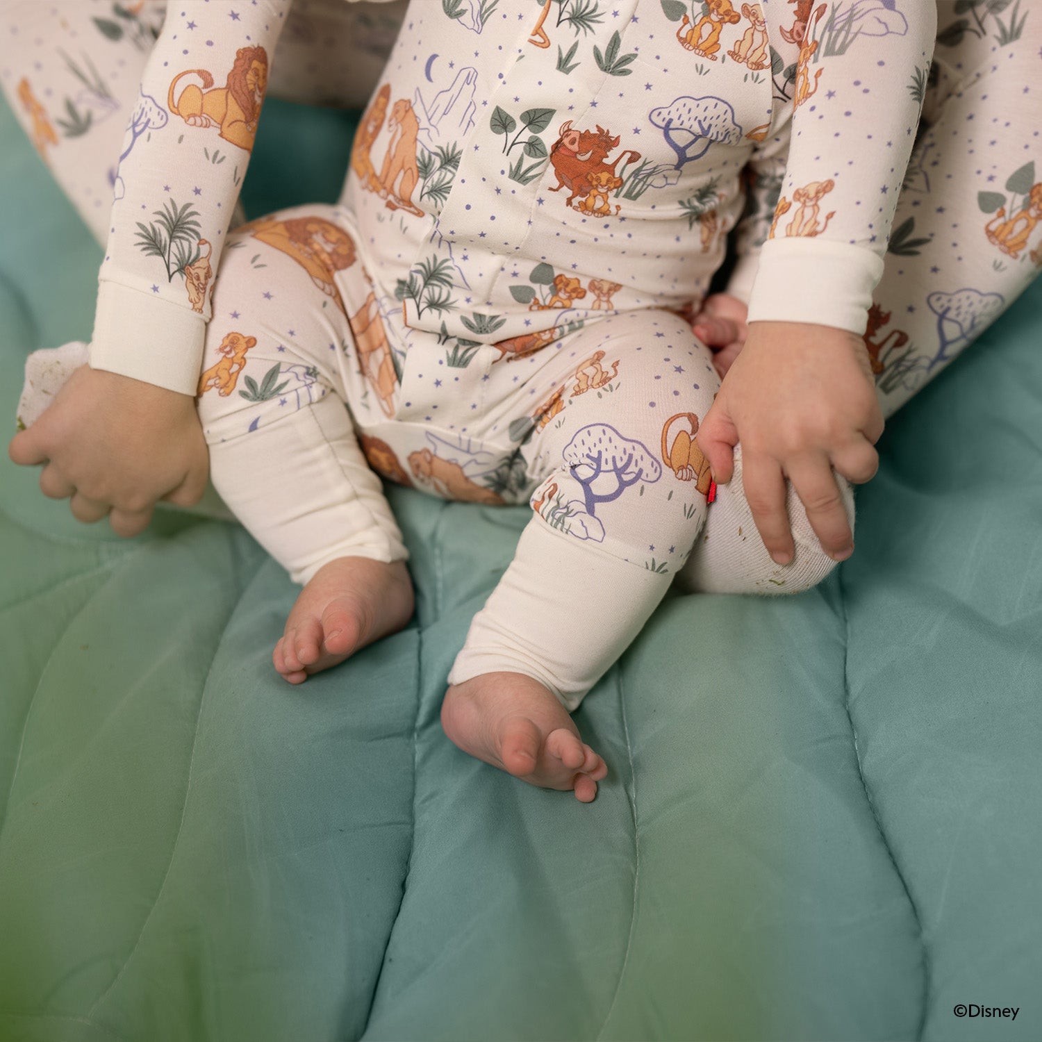 Close up photo of the legs of a baby who is wearing A cream-colored long-sleeve coverall featuring a repeating pattern of Disney Pumbaa, Simba, and Nala among savanna trees and blue stars by Magnetic Me.