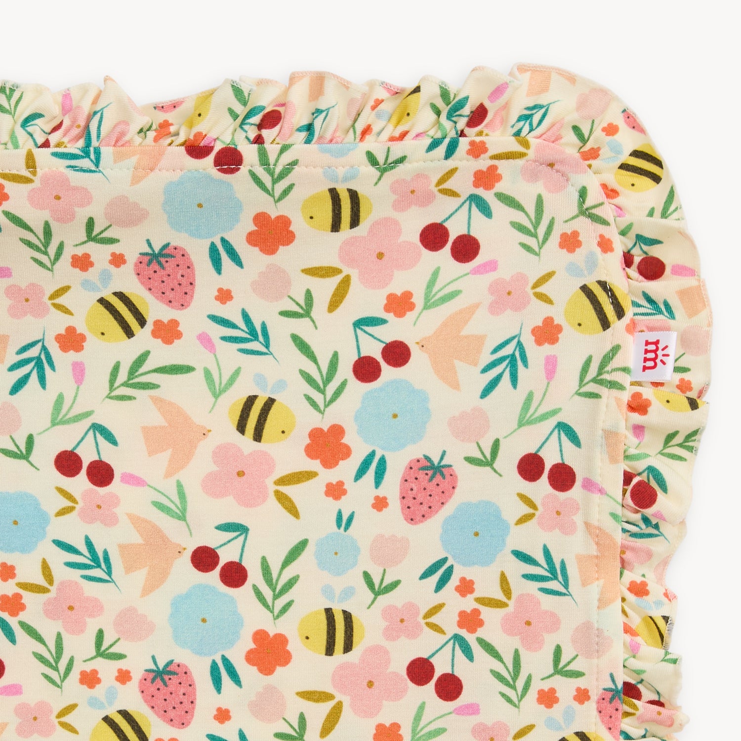 A cream-colored baby blanket featuring a whimsical garden print of bees, strawberries, cherries, and flowers with a ruffled edge, by Magnetic Me.