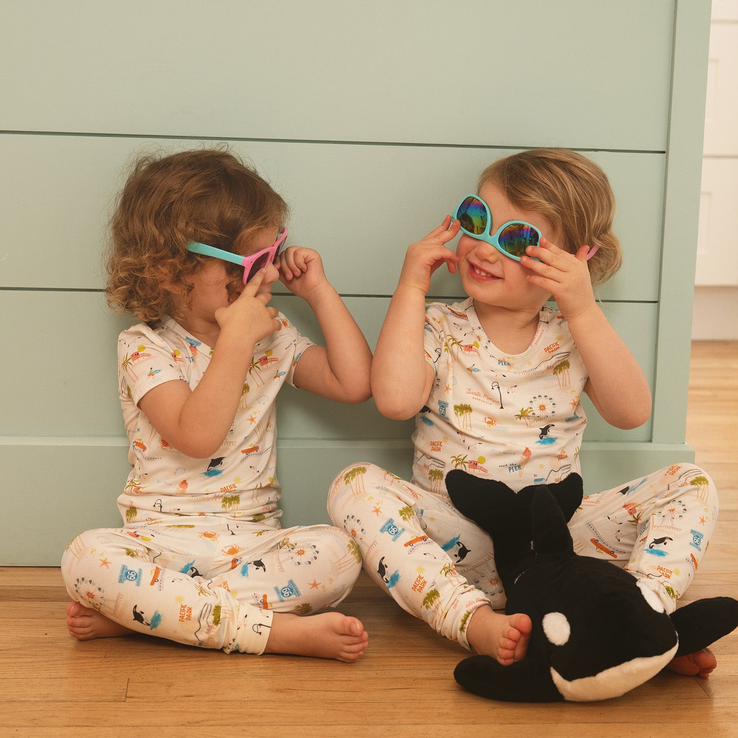 Two children wearing colorful sunglasses sitting on a wooden floor with a plush toy wearing A sleepwear set includes a short sleeve top and matching long pants adorned with the detailed Santa Monica coastline and amusement park print by Magnetic Me.
