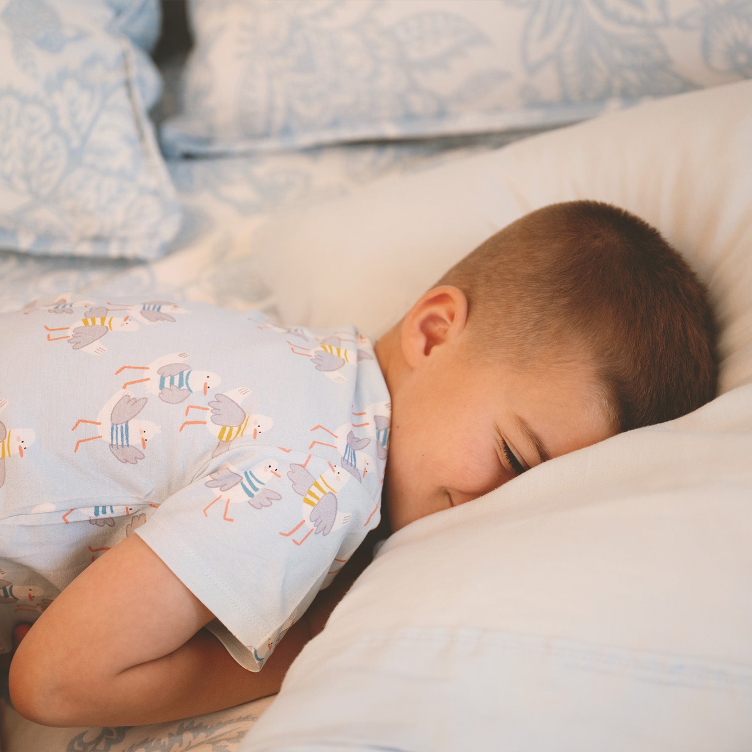 Child sleeping in bed with patterned pajamas wearing A two-piece children's pajama set including a short-sleeved top and matching shorts in light blue with an all-over seagull sweater print, by Magnetic Me.