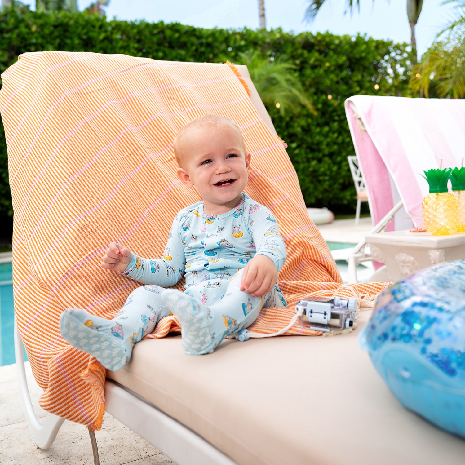 Baby sitting on a lounge chair by a poolside with a colorful towel and pool toy while wearing A long-sleeve light blue footie featuring a playful print of raccoons lounging on colorful pool floats and snorkeling, by Magnetic Me.