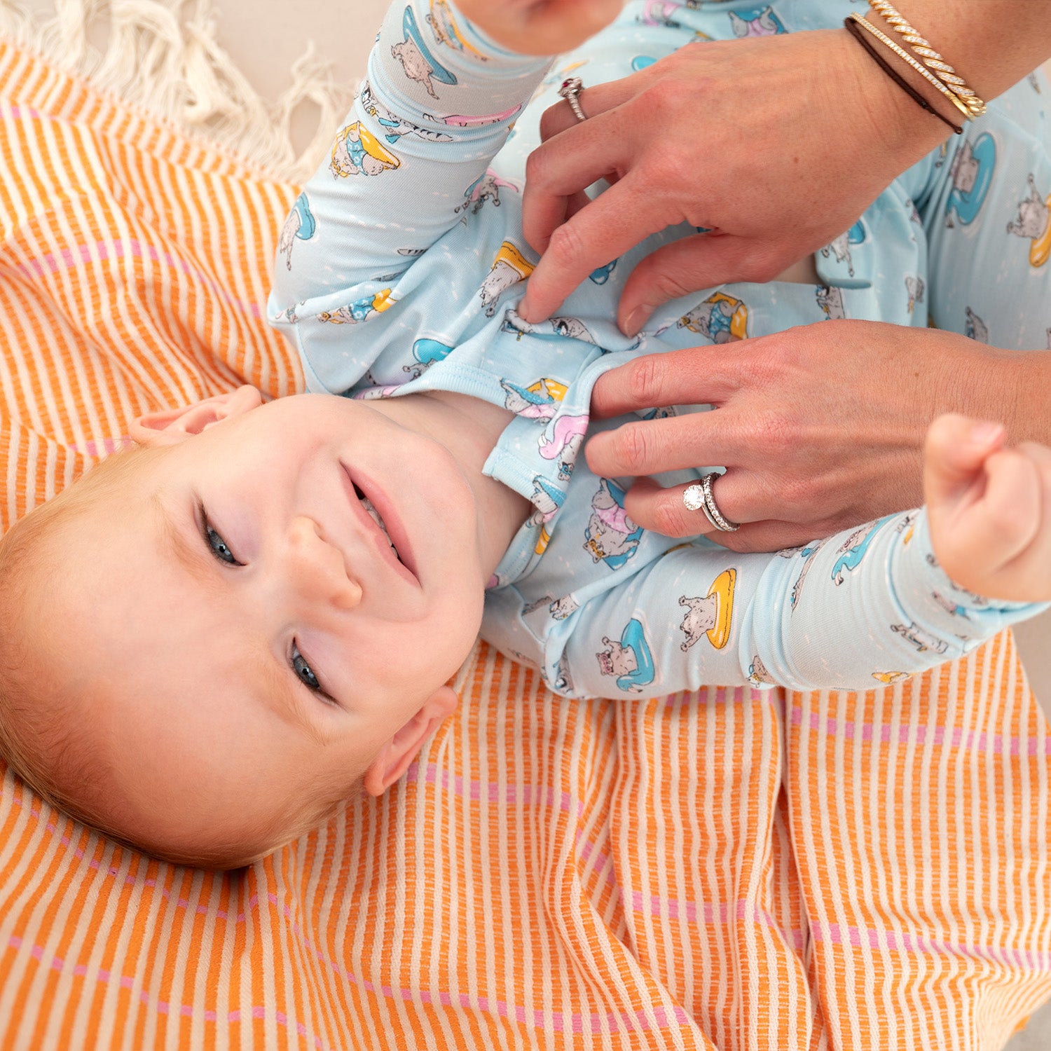 Baby wearing lying on an orange and pink checkered blanket and wearing A long-sleeve light blue footie featuring a playful print of raccoons lounging on colorful pool floats and snorkeling, by Magnetic Me.