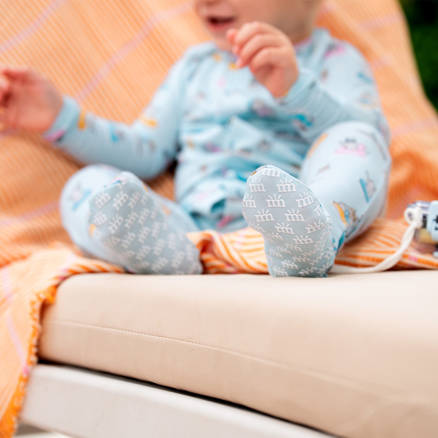Baby on an orange blanket wearing A long-sleeve light blue footie featuring a playful print of raccoons lounging on colorful pool floats and snorkeling, by Magnetic Me.