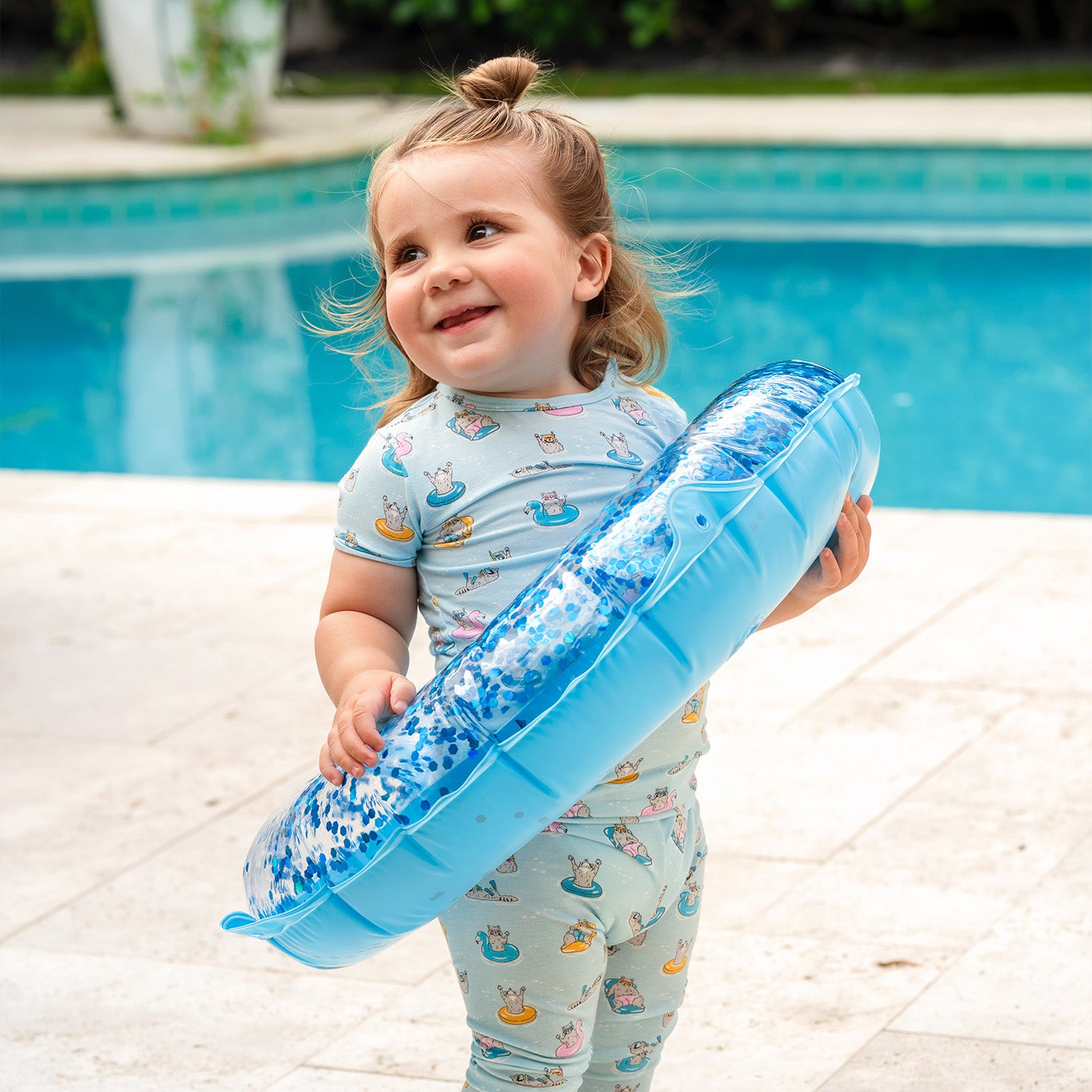 Child holding a blue inflatable ring by a pool and wearing A two-piece pajama set in light blue featuring a repeating print of raccoons on bright pool floats and wearing goggles, by Magnetic Me.