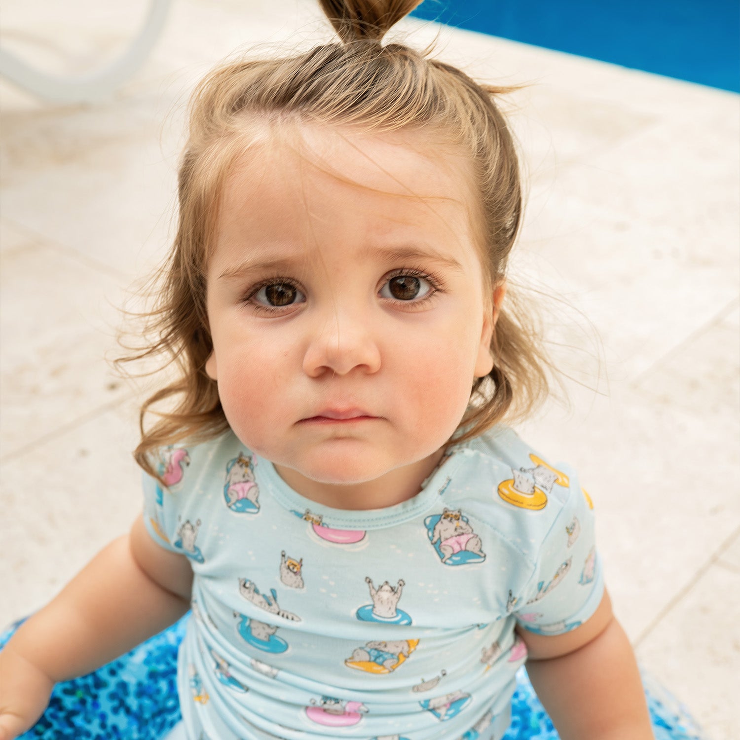 Young child by a pool wearing A two-piece pajama set in light blue featuring a repeating print of raccoons on bright pool floats and wearing goggles, by Magnetic Me.