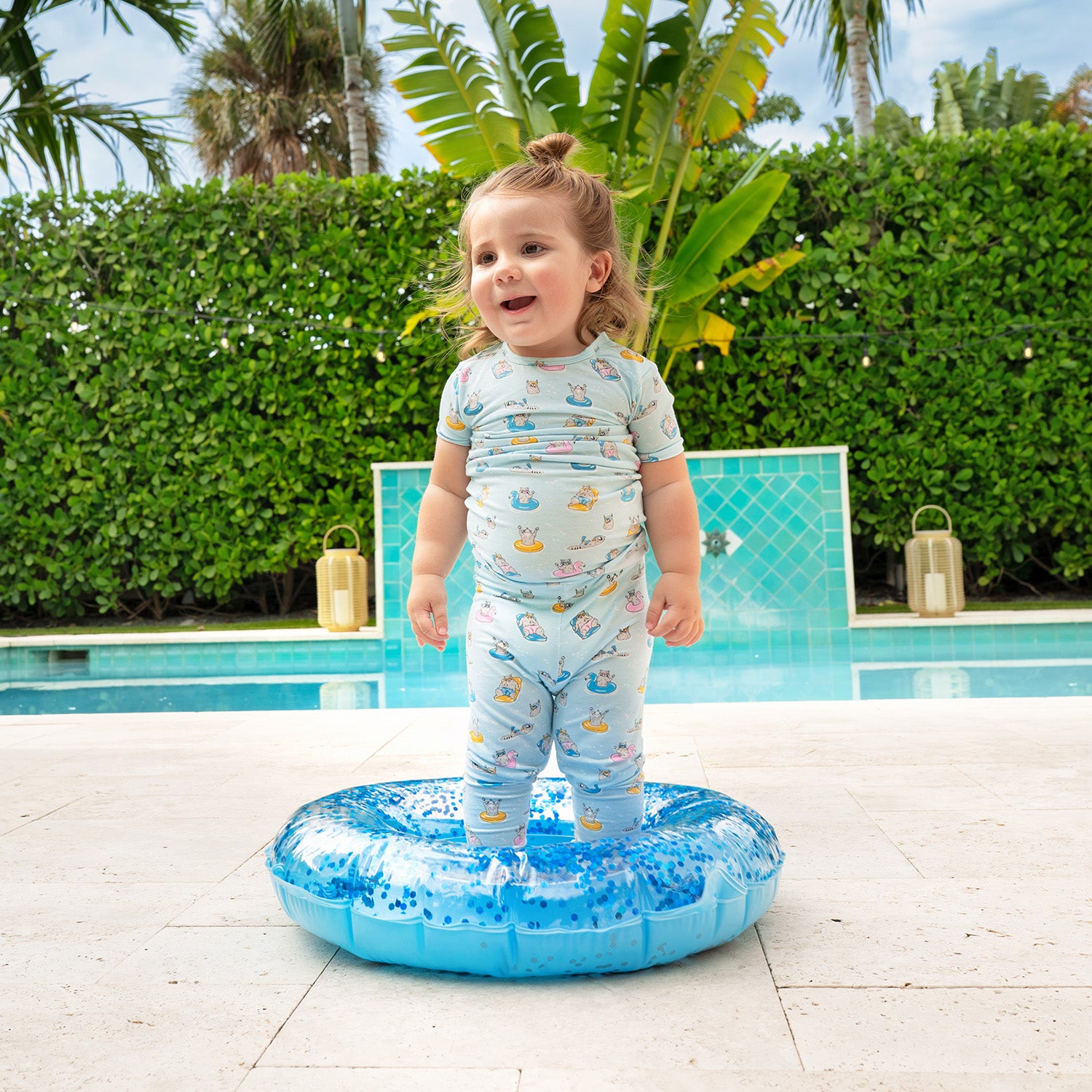 Child standing on a small inflatable pool toy by a pool and wearing A two-piece pajama set in light blue featuring a repeating print of raccoons on bright pool floats and wearing goggles, by Magnetic Me.