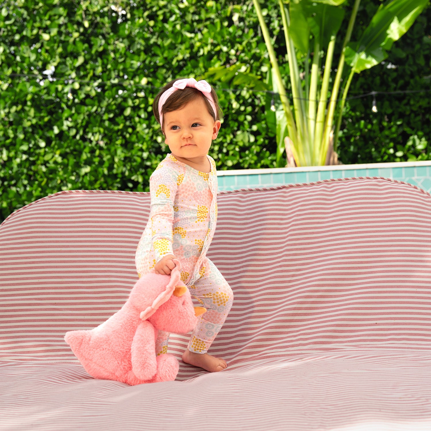 Baby holding a pink plush toy, sitting on a striped chair cover while wearing A long-sleeve convertible coverall with ruffle detailing and a multi-colored pastel floral pattern, by Magnetic Me.