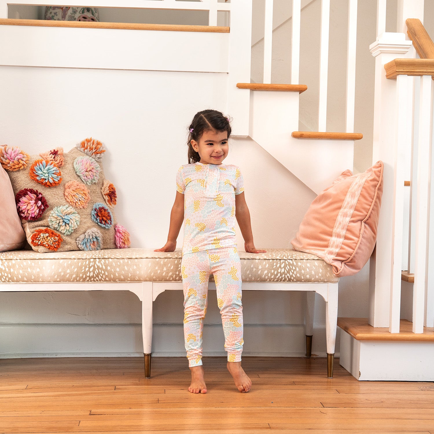 Child by a staircase with decorative pillows wearing A two-piece pajama set featuring a short-sleeve ruffle-trim top and matching pants in a bright pastel spring floral print, by Magnetic Me.