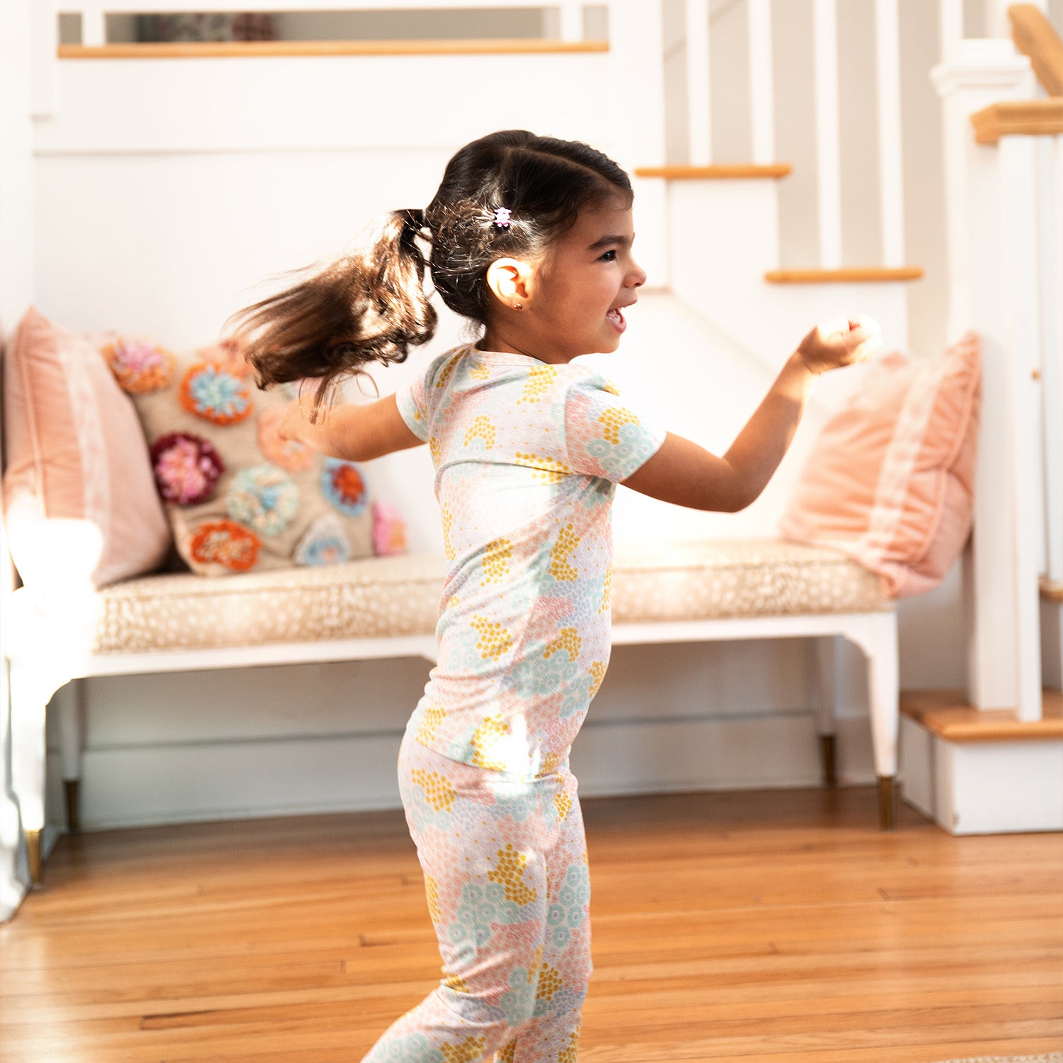Child running around a room with a couch and pillows wearing A two-piece pajama set featuring a short-sleeve ruffle-trim top and matching pants in a bright pastel spring floral print, by Magnetic Me.