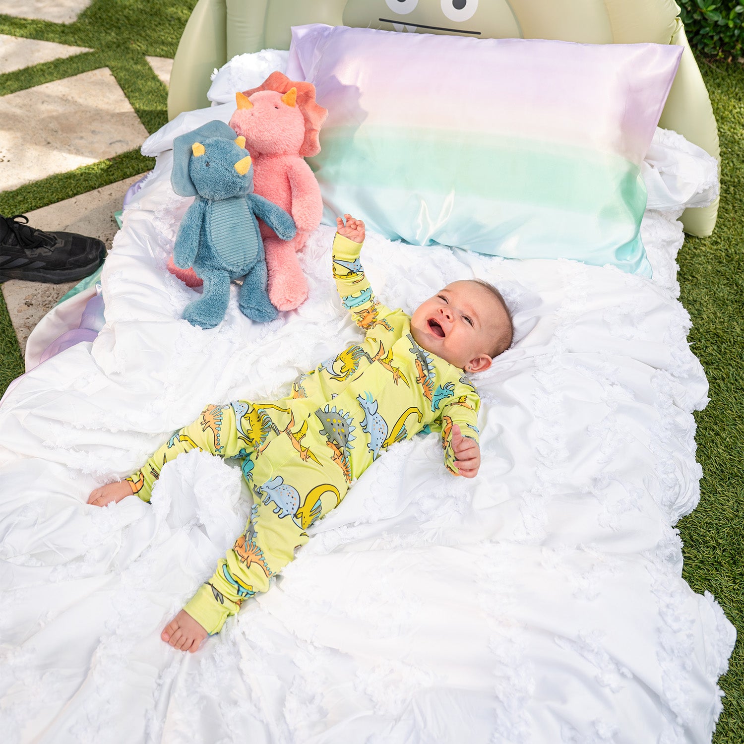 Baby on a white blanket with two plush toys, surrounded by green grass wearing A lime green long-sleeve convertible coverall with a playful multi-colored dinosaur pattern and ribbed cuffs, by Magnetic Me.