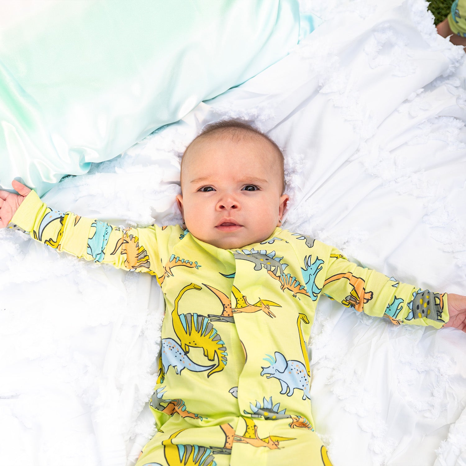 Baby lying on a white blanket wearing A lime green long-sleeve convertible coverall with a playful multi-colored dinosaur pattern and ribbed cuffs, by Magnetic Me.