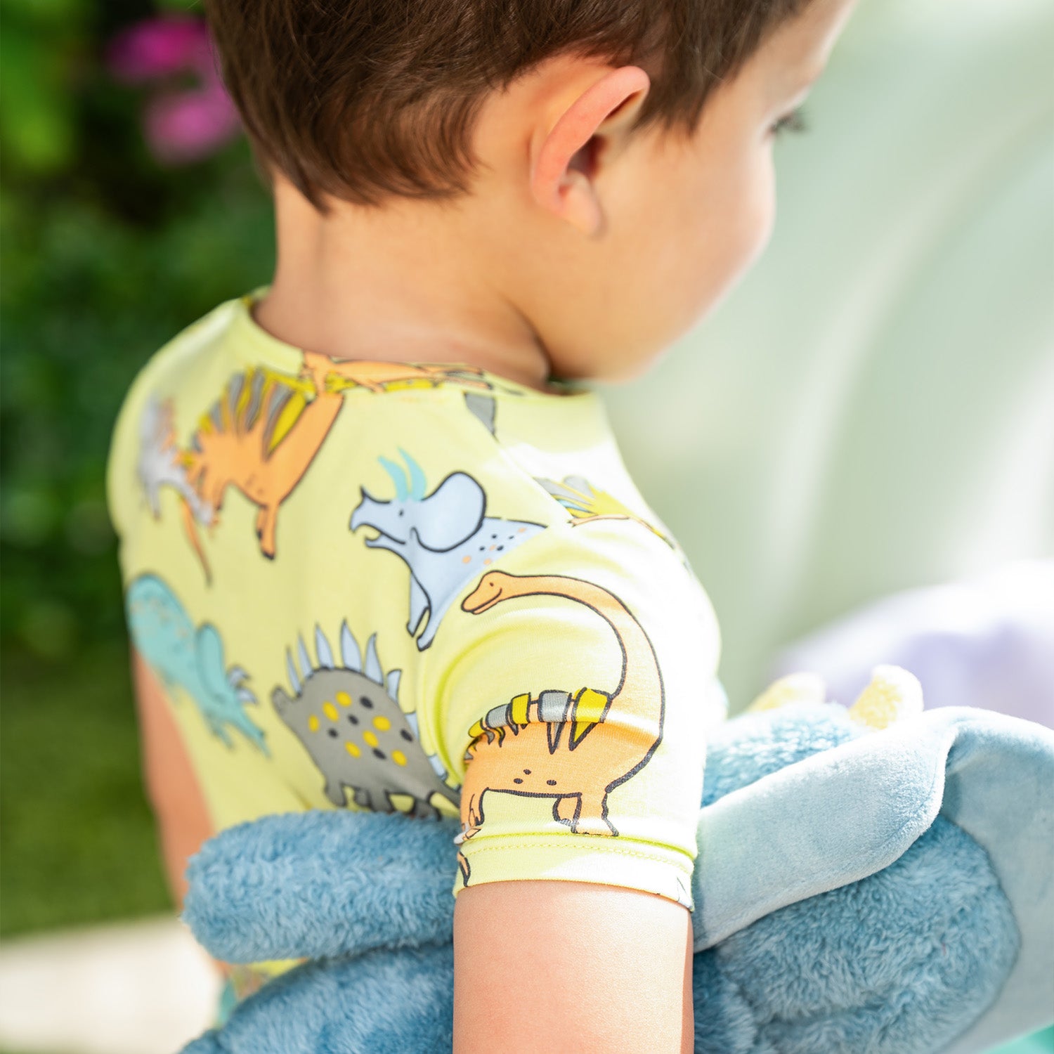 Child holding a blue plush toy outdoors wearing A two-piece pajama set with a short-sleeve top and long pants in lime green fabric covered in a whimsical dinosaur print, by Magnetic Me.