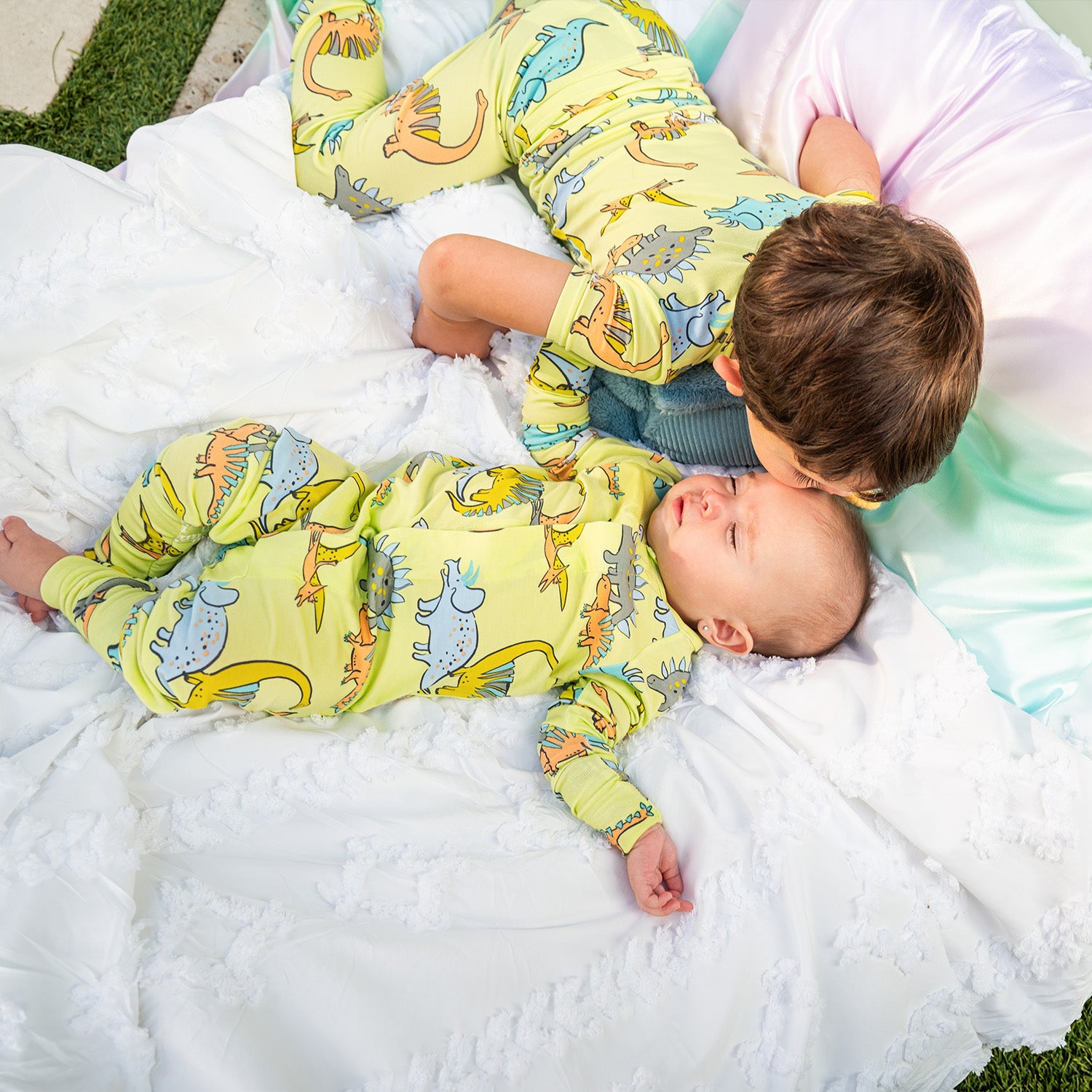 Two children wearing matching Roar 
& Explore Nightwear lying on a white blanket.