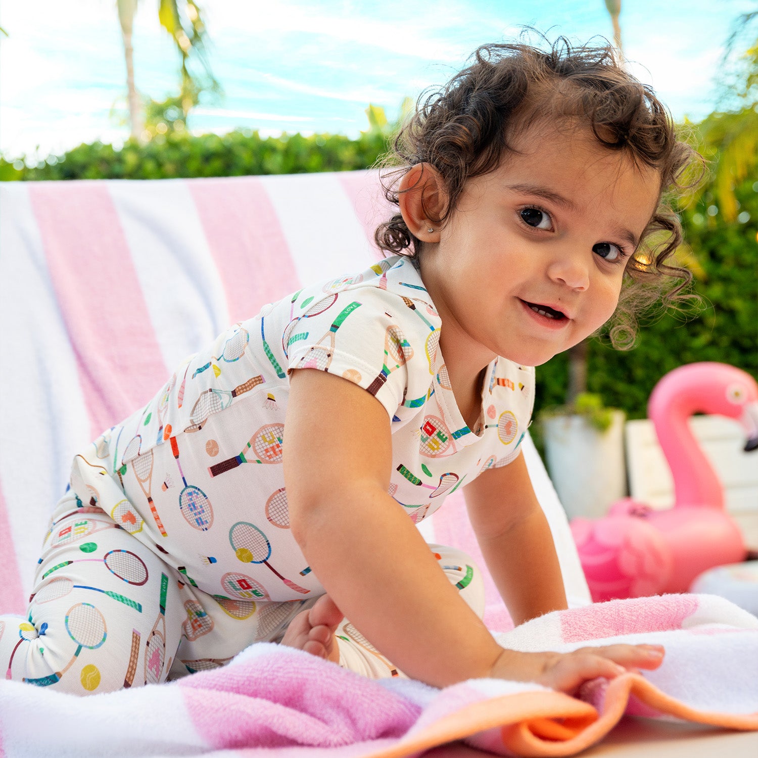 Child sitting on a colorful blanket with a pink flamingo toy wearing A two-piece pajama set with a cream short-sleeve top and long pants covered in a whimsical tennis and badminton equipment print, by Magnetic Me.