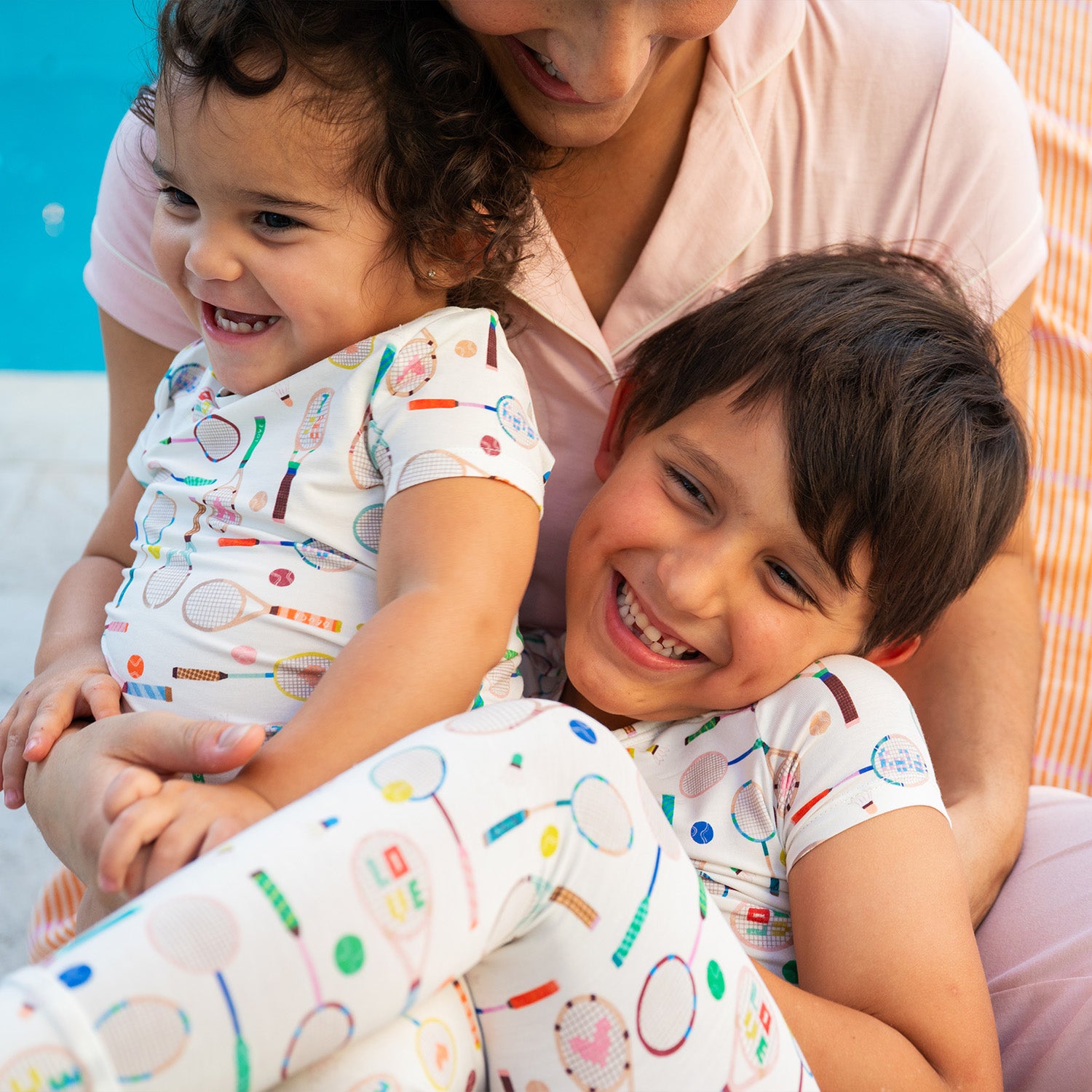 Woman holding two children wearing matching two-piece pajama set with a cream short-sleeve top and long pants covered in a whimsical tennis and badminton equipment print, by Magnetic Me.