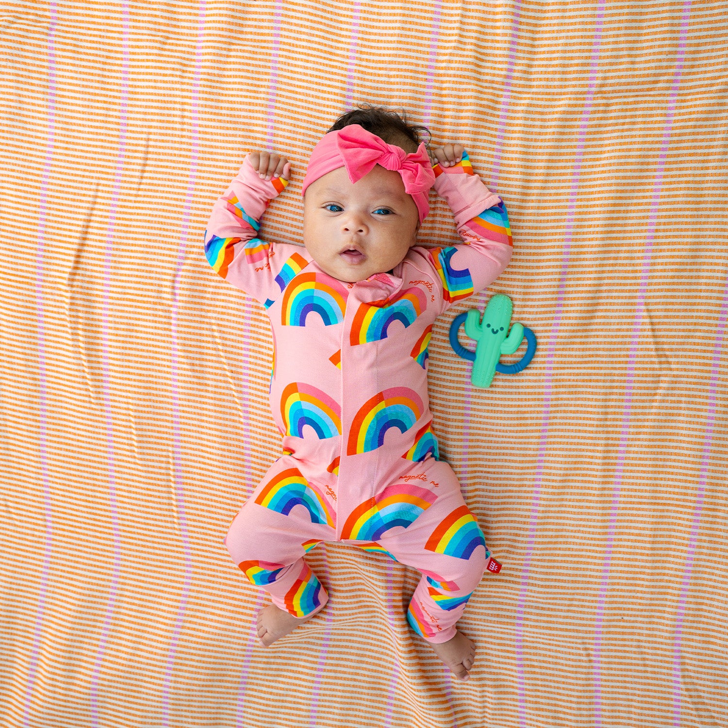 Baby lying on a striped blanket wearing A pink long-sleeve convertible coverall decorated with a colorful all-over rainbow print and ribbed cuffs, by Magnetic Me.