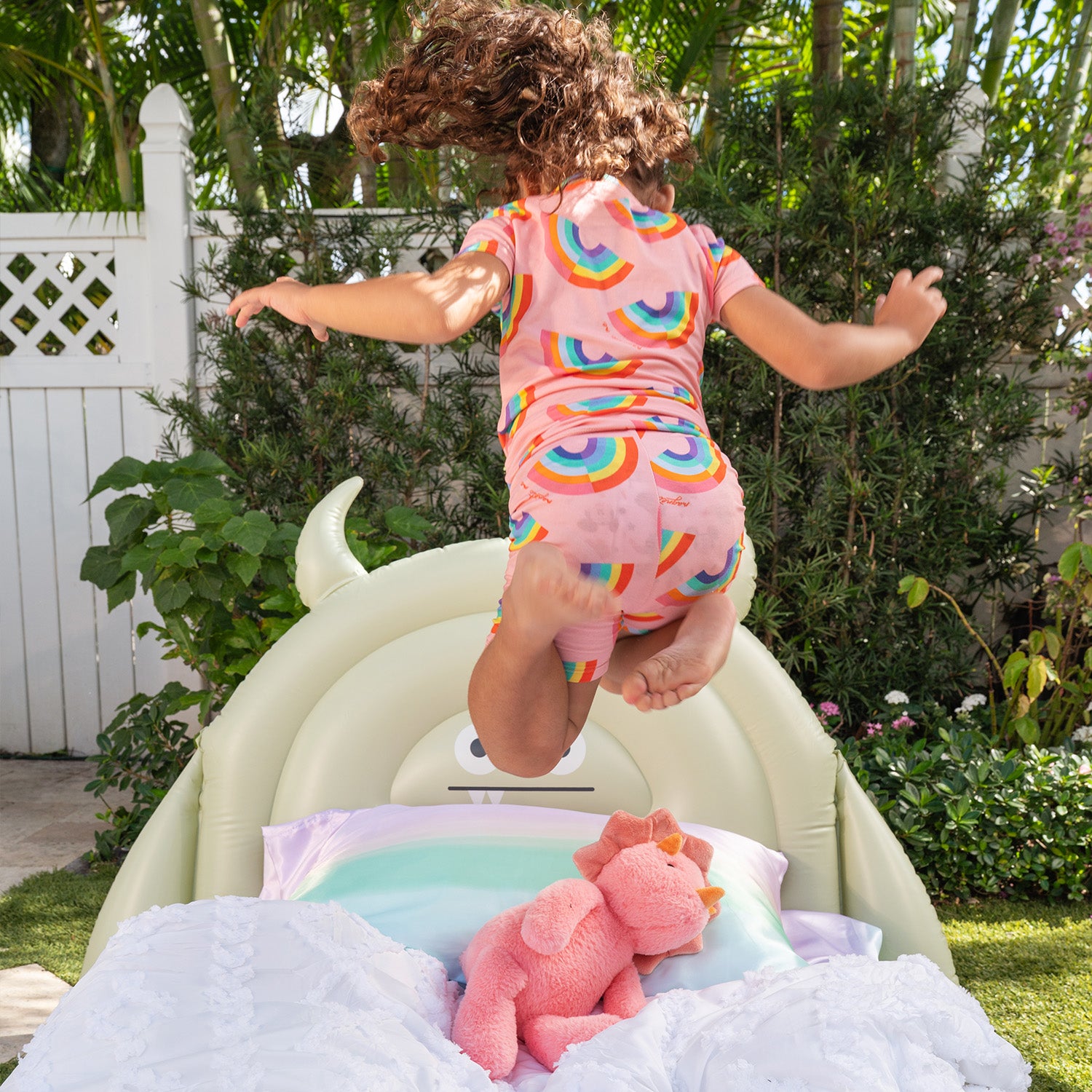 Child in a colorful outfit jumping on a bed and wearing A two-piece pajama set with a short-sleeve top and matching shorts in pink fabric featuring a bright, repeating rainbow pattern, by Magnetic Me.