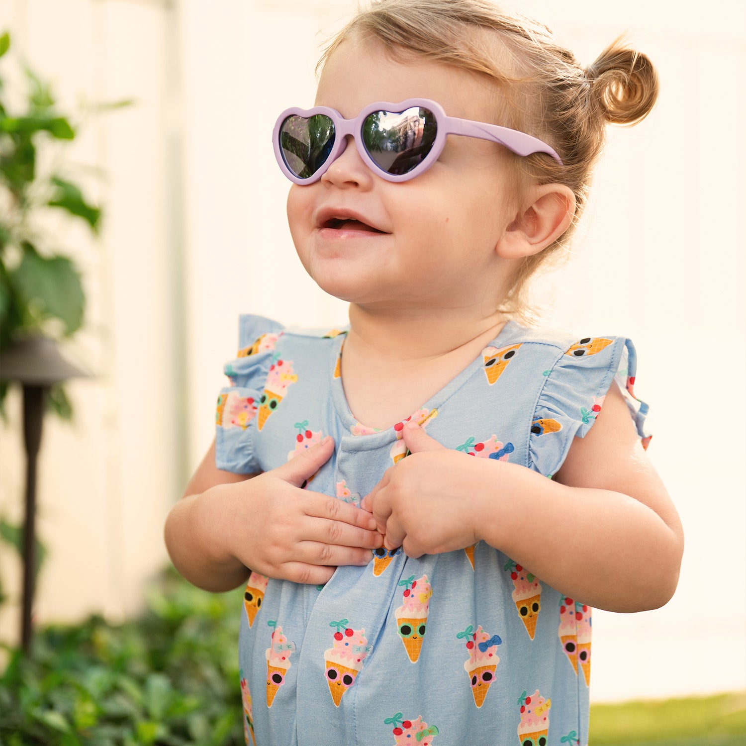 Child wearing heart-shaped sunglasses and A cornflower blue sleeveless romper with ruffled flutter sleeves and cinched leg openings, featuring a charming ice cream cone character print, by Magnetic Me.