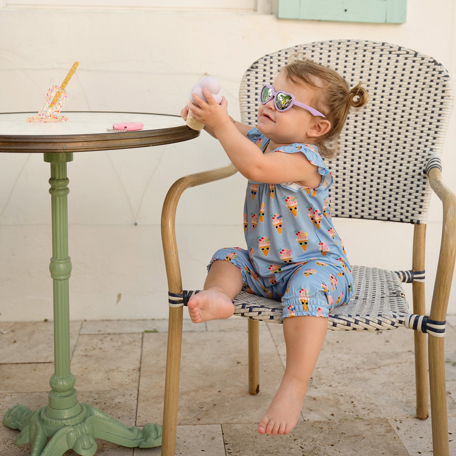 Child sitting on a chair outdoors with ice cream wearing heart shaped sunglasses and A cornflower blue sleeveless romper with ruffled flutter sleeves and cinched leg openings, featuring a charming ice cream cone character print, by Magnetic Me.