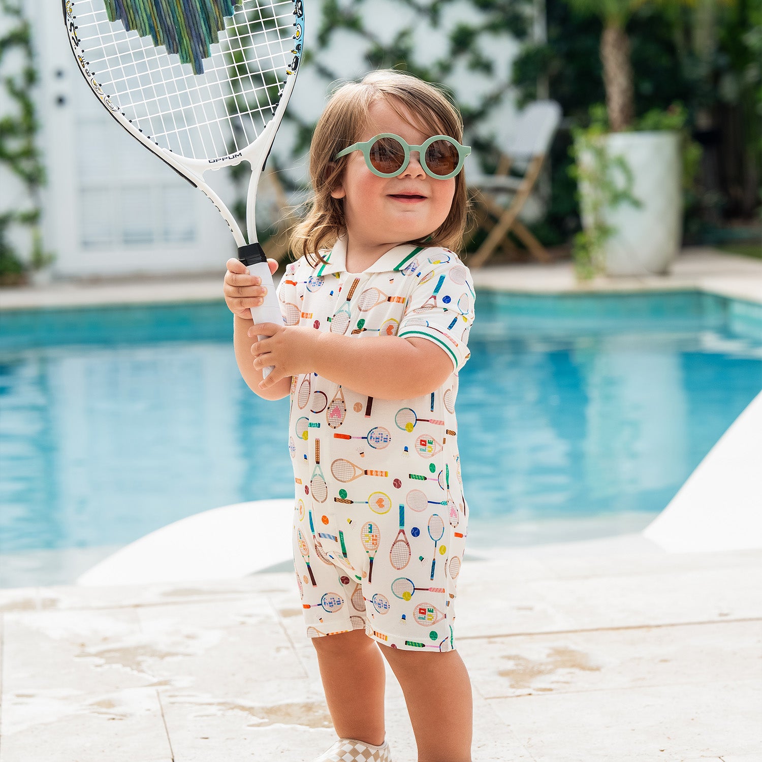 Child holding a tennis racket by a pool and wearing A cream-colored short-sleeve romper with a green-trimmed polo collar, featuring a repeating print of tennis rackets, shuttlecocks, and colorful sports balls, by Magnetic Me.