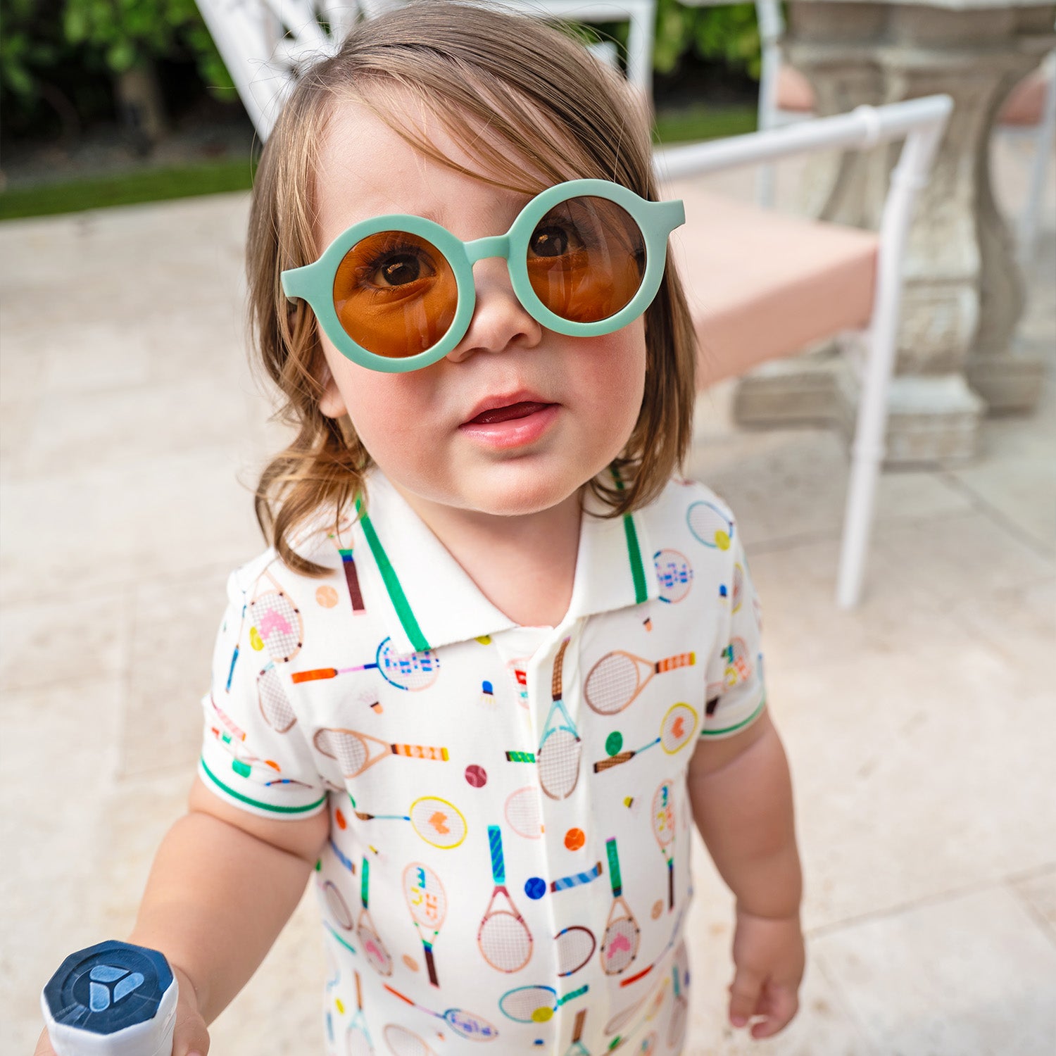 Child wearing colorful sunglasses and A cream-colored short-sleeve romper with a green-trimmed polo collar, featuring a repeating print of tennis rackets, shuttlecocks, and colorful sports balls, by Magnetic Me.
