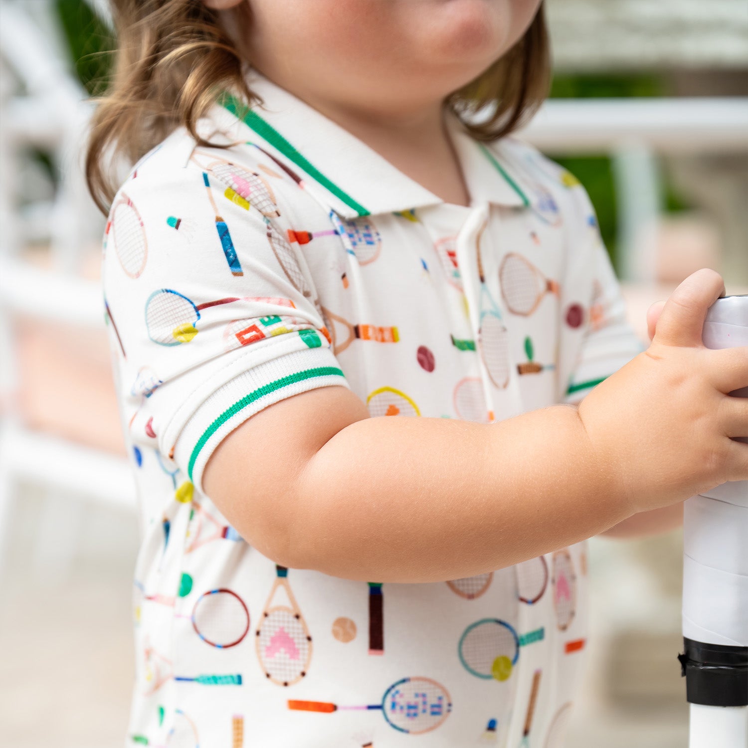 Close up photo of the child wearing A cream-colored short-sleeve romper with a green-trimmed polo collar, featuring a repeating print of tennis rackets, shuttlecocks, and colorful sports balls, by Magnetic Me.