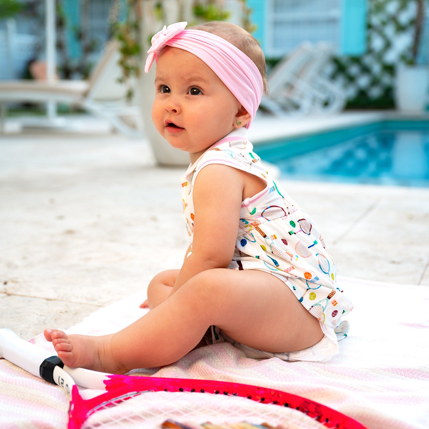 Baby sitting on a towel by a pool wearing a pink headband and A sleeveless cream-colored dress with a pink-trimmed polo collar and an integrated bodysuit, featuring a colorful print of tennis rackets, balls, and shuttlecocks, by Magnetic Me.