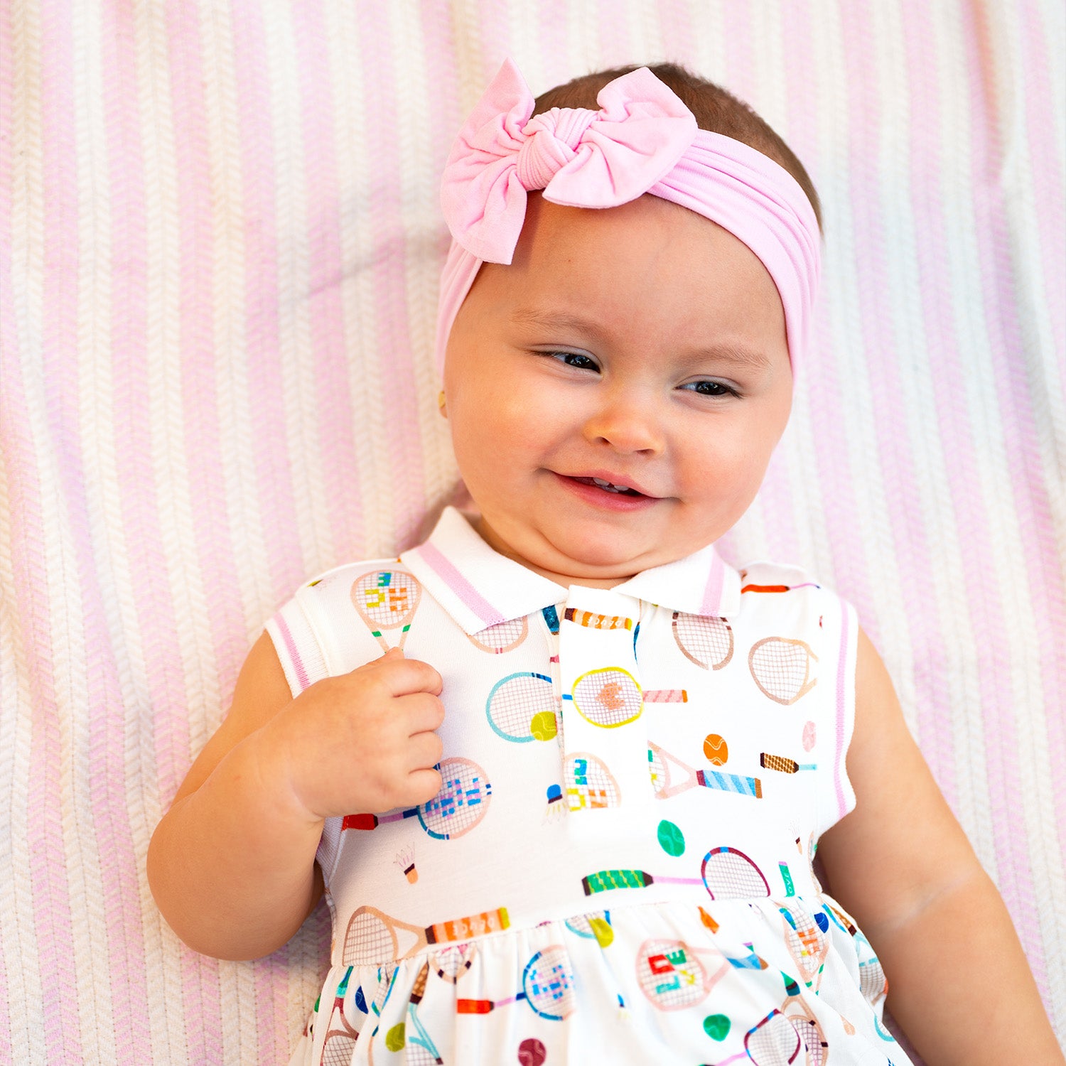 Baby wearing a pink headband against a pink and A sleeveless cream-colored dress with a pink-trimmed polo collar and an integrated bodysuit, featuring a colorful print of tennis rackets, balls, and shuttlecocks, by Magnetic Me.