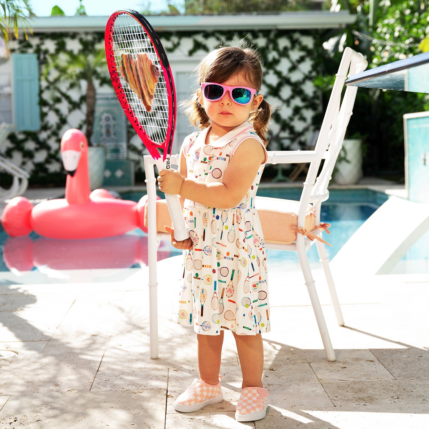 Child holding a tennis racket by a poolside with a flamingo floaty in the background while wearing A cream sleeveless polo dress with a grey and white striped collar, decorated with a whimsical pattern of multi-colored tennis and badminton equipment, by Magnetic Me.